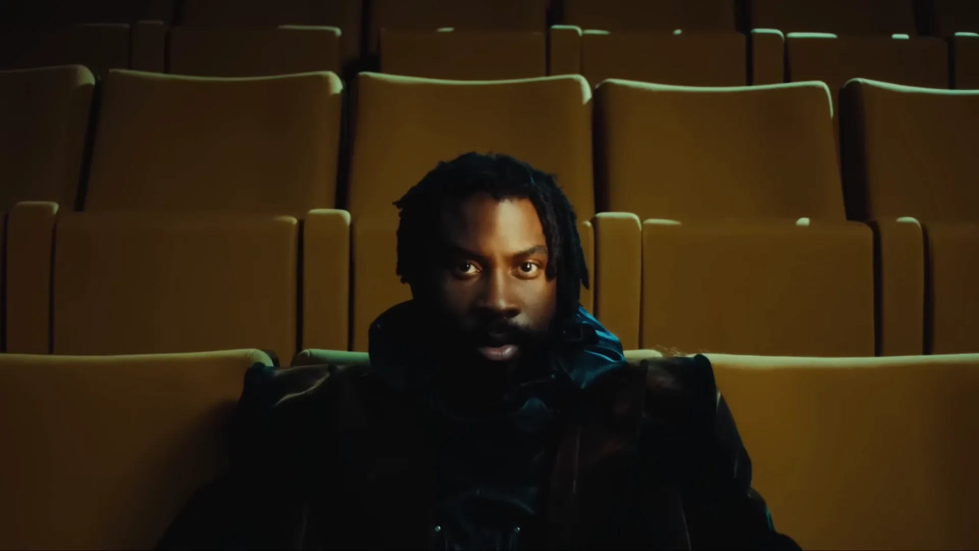 Damso assis seul dans une salle de cinéma aux fauteuils jaunes, dreadlocks et manteau sombre, regard face caméra, dans le clip « Laisse-moi tranquille. »