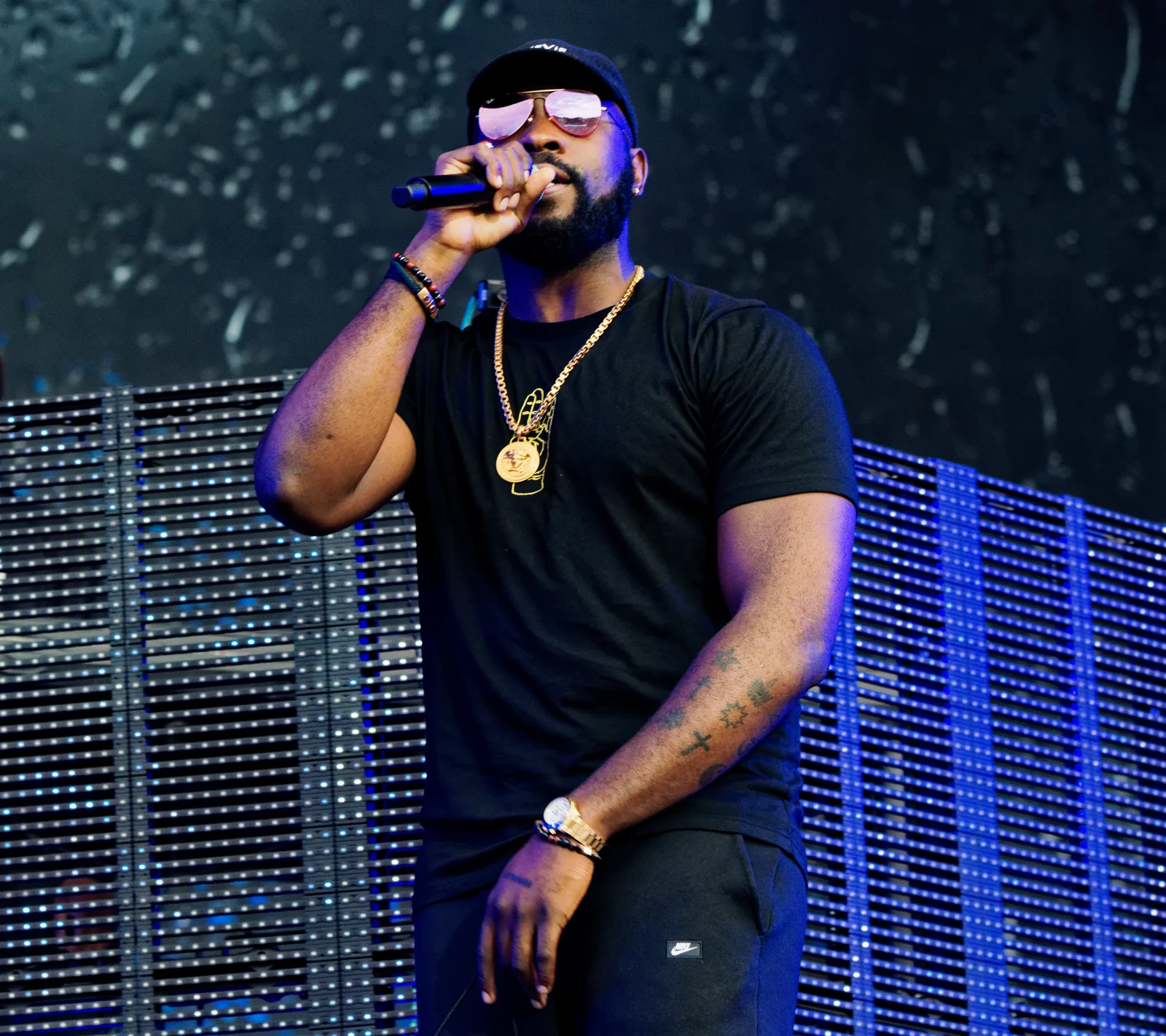 Damso rappe micro en main, casquette noire et lunettes de soleil, en tee-shirt noir et chaîne dorée, devant un mur de lumières bleues.