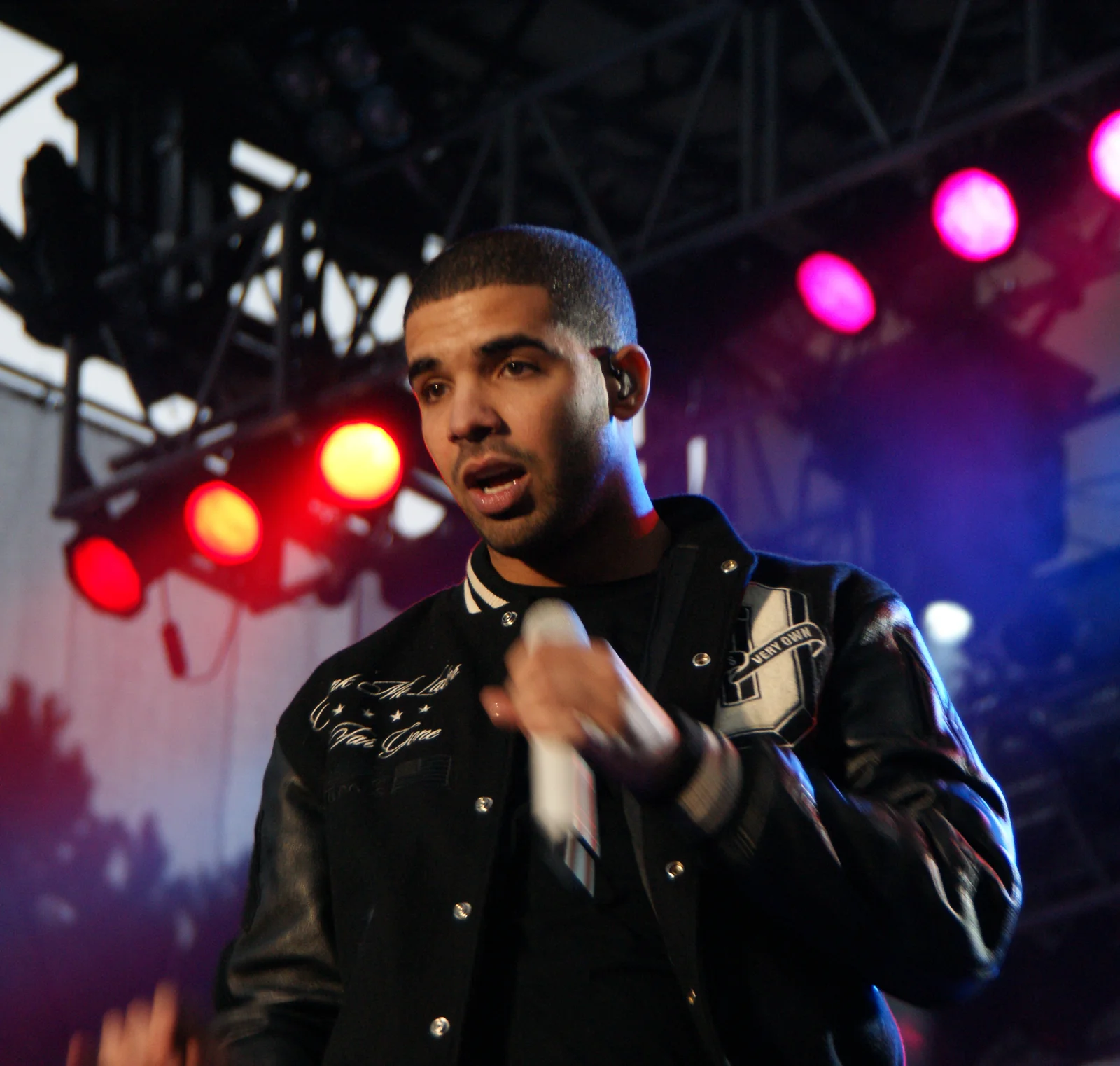 Drake chante micro en main, en blouson de type universitaire noir, devant les projecteurs rouges et roses d’une scène de festival.