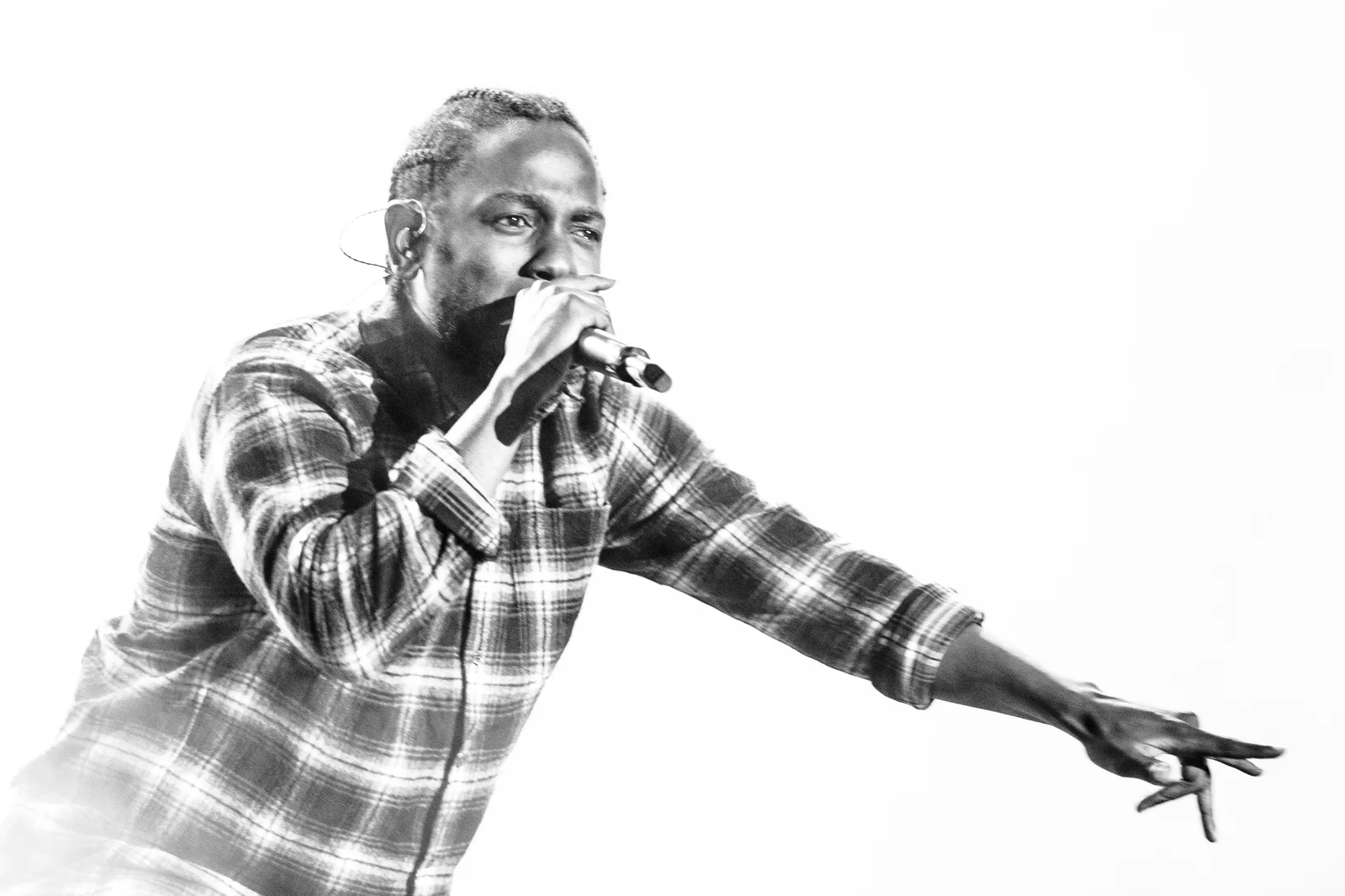 Photo en noir et blanc : Kendrick Lamar rappe micro en main, en chemise à carreaux, bras tendu vers le public, sur fond blanc lumineux.