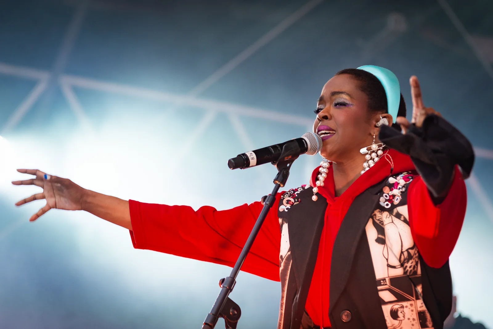 Lauryn Hill chante au micro sur pied, bras tendu vers le public, en sweat rouge et veste imprimée, sous les projecteurs d’une scène en plein air.
