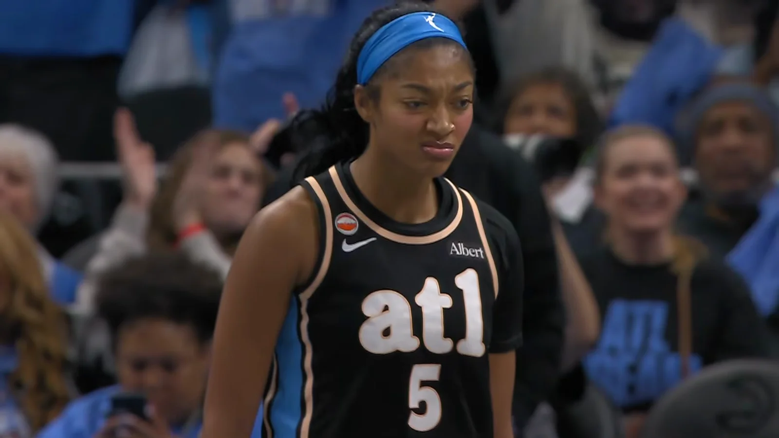 Angel Reese, bandeau bleu et maillot noir « atl 5 » de l’Atlanta Dream, fixe l’objectif la mâchoire serrée, devant des supporters debout en tribune.