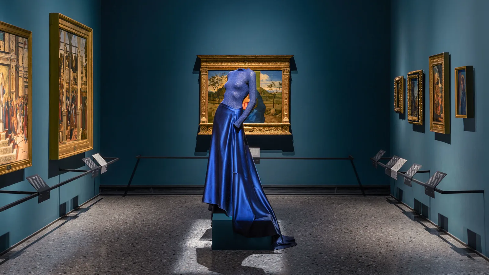 Une robe Giorgio Armani bleu roi, au bustier scintillant et à la longue jupe de satin, exposée au centre d’une salle aux murs bleu canard de la Pinacothèque de Brera, entre des tableaux anciens à cadres dorés.