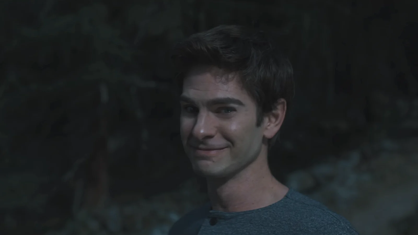 Andrew Garfield, dans le rôle de Sam Altman, sourit en tee-shirt gris dans un sous-bois à la tombée du jour, plan tiré de la bande-annonce d’Artificial.