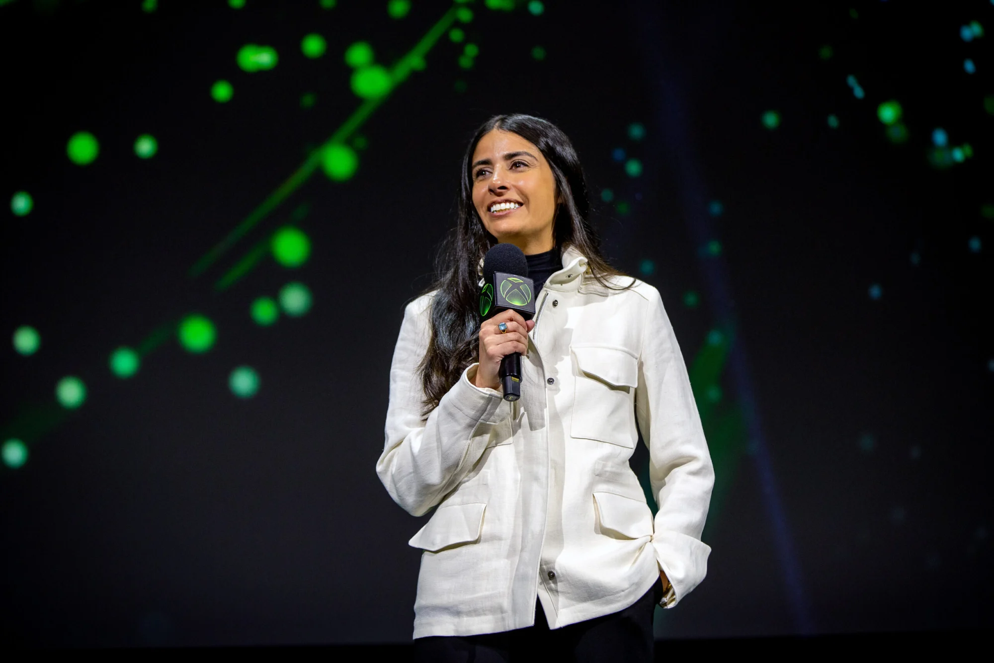 Asha Sharma, PDG de Xbox, sourit sur scène en veste blanche, un micro au logo Xbox à la main, devant un écran noir parsemé de points lumineux verts.