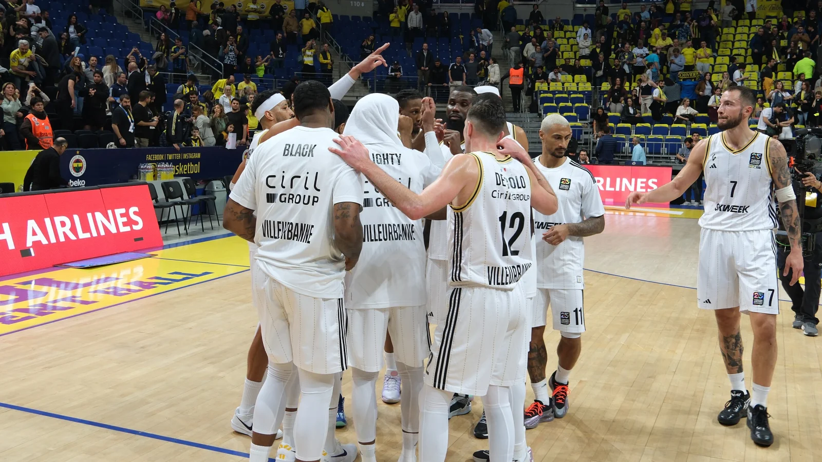Les joueurs de l’ASVEL, en maillots blancs floqués Villeurbanne, réunis en cercle sur le parquet du Fenerbahçe à Istanbul après un match d’EuroLeague, devant des tribunes jaunes et bleues.