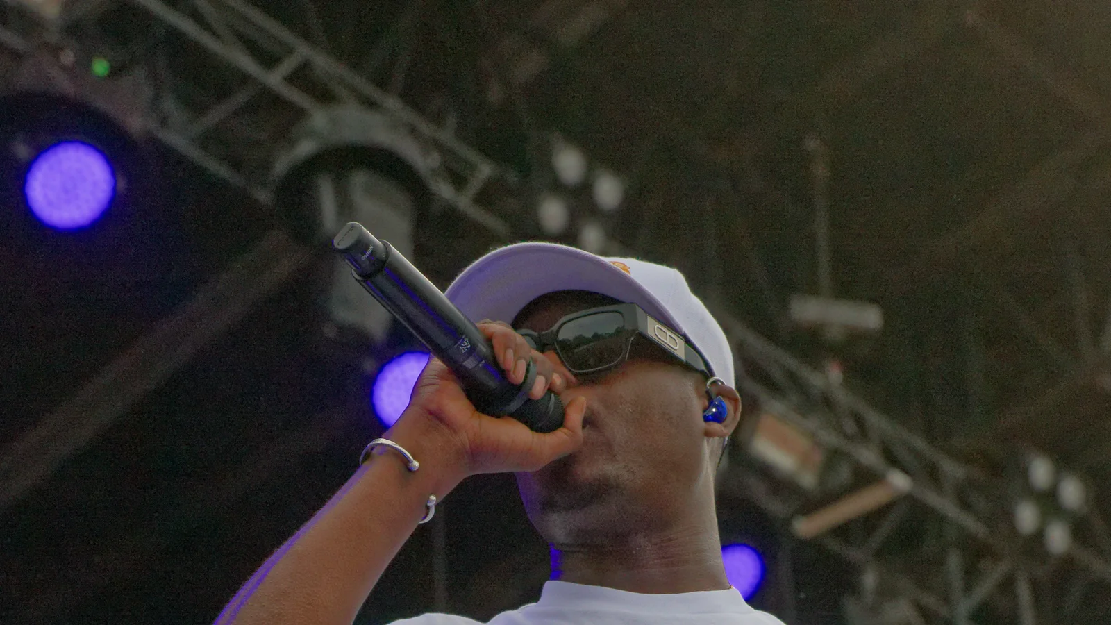 Le rappeur Ninho sur scène, casquette blanche et lunettes de soleil, chante au micro sous les structures d’éclairage du festival des Vieilles Charrues.