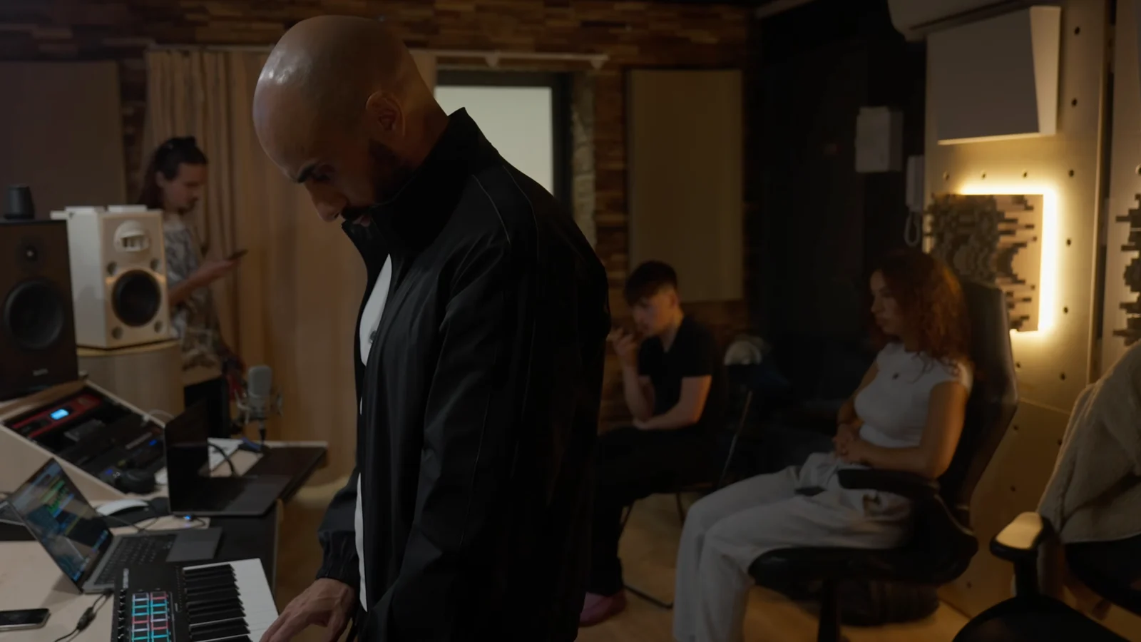 Dans un studio aux murs de briques et de panneaux acoustiques éclairés, le producteur Tarik Azzouz, debout en veste noire, joue sur un clavier maître à pads lumineux, entre console, ordinateurs portables, micro et enceintes ; au fond, trois personnes de la session l'écoutent.