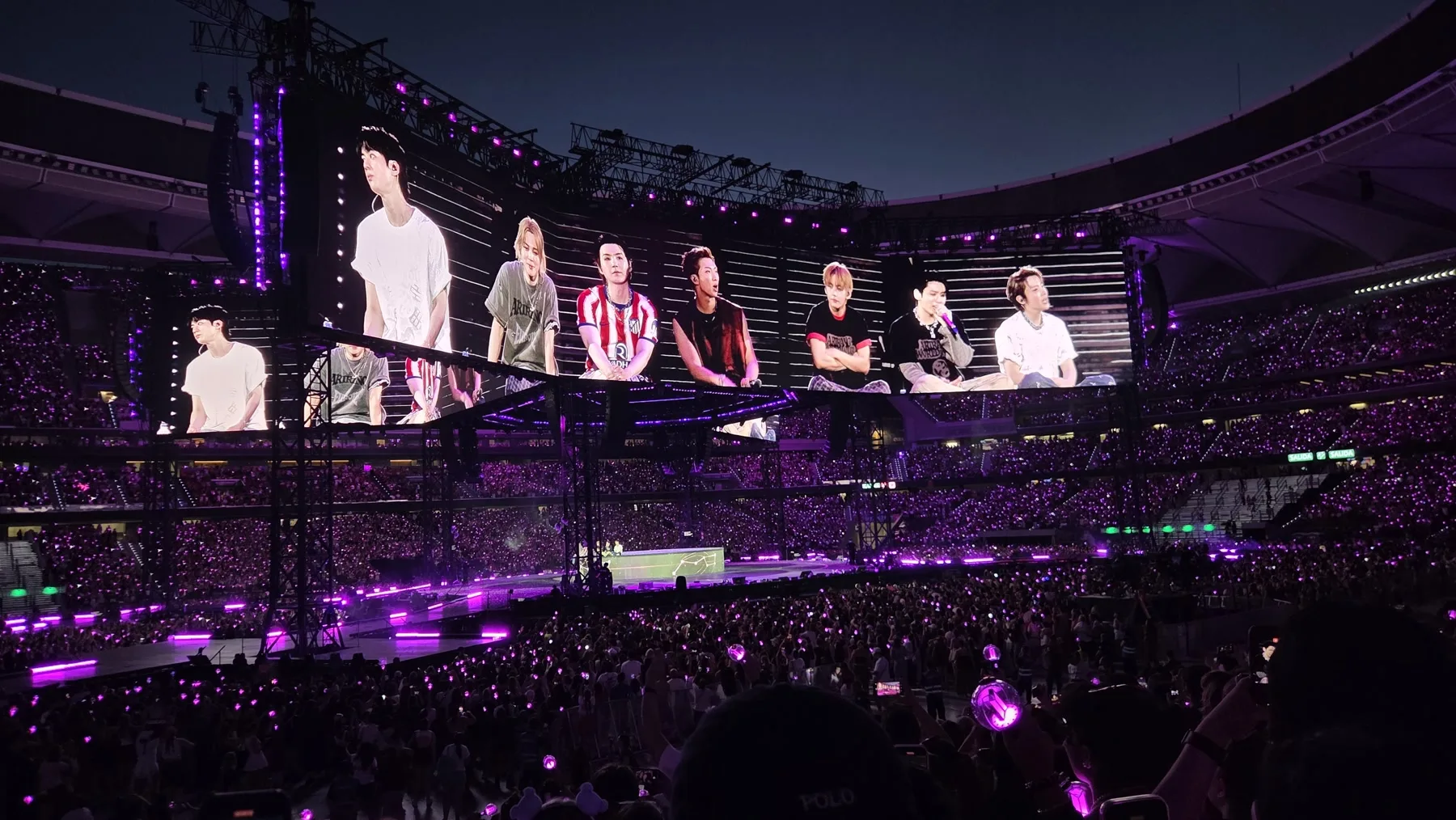 Les membres de BTS sur l’écran géant d’un stade éclairé en violet, pendant la tournée Arirang à Madrid.
