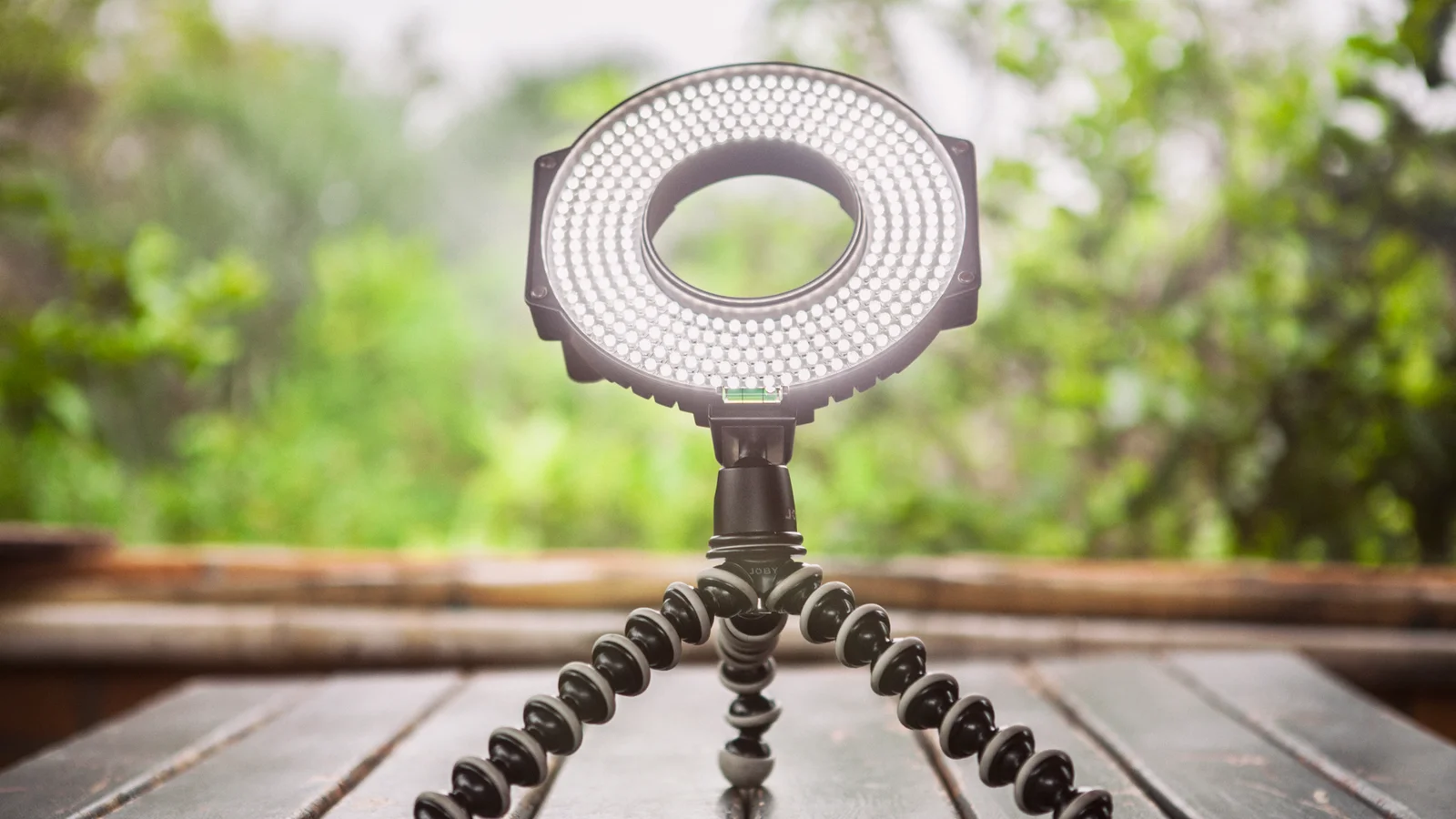 Un anneau lumineux LED allumé, fixé sur un petit trépied à pieds articulés, posé sur une table en bois devant un fond de verdure flou.