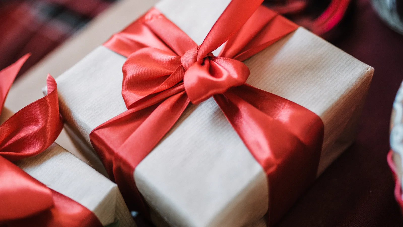 Un paquet cadeau emballé de papier kraft clair et noué d’un large ruban de satin rouge, posé sur une table sombre à côté d’un second paquet identique.