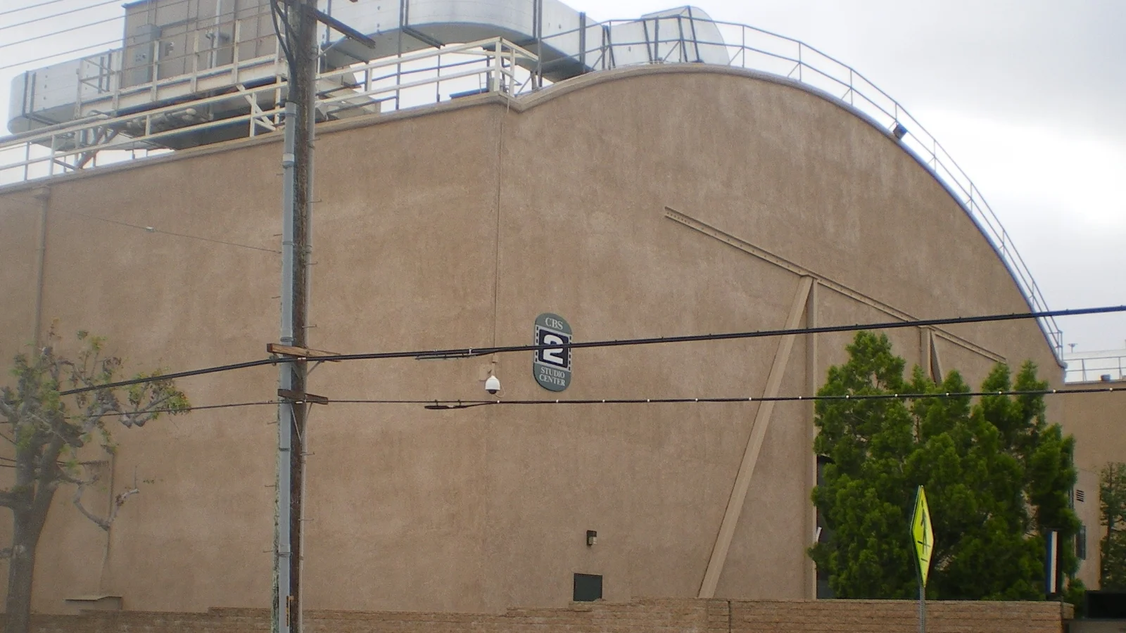 Façade beige et cintrée d'un plateau de tournage, marquée d'un grand chiffre 2, derrière un mur de briques et des poteaux électriques.
