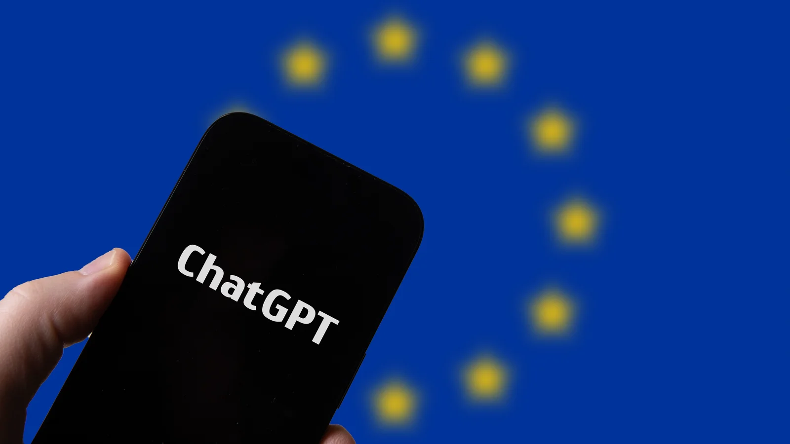 Une main tient un smartphone dont l’écran noir affiche le nom ChatGPT, devant le drapeau bleu aux étoiles jaunes de l’Union européenne, flou à l’arrière-plan.
