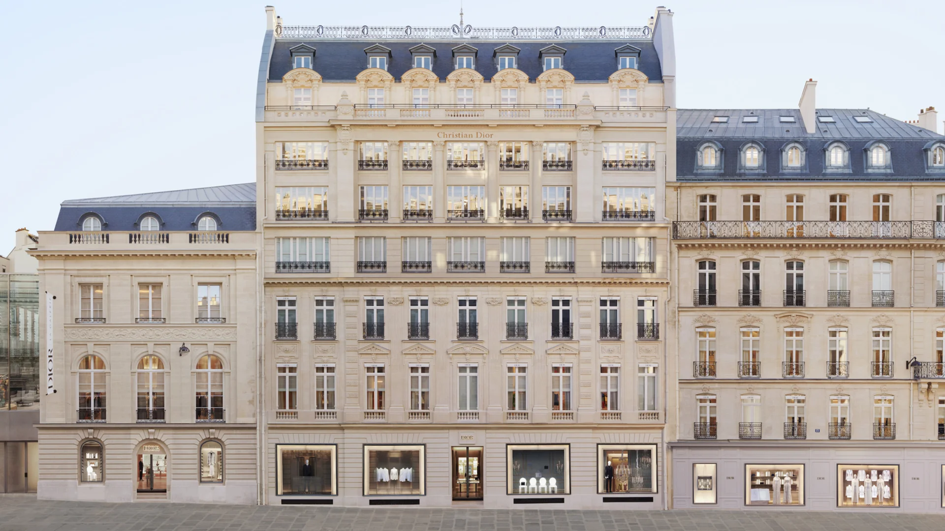 La façade en pierre de taille de la maison Dior au 30, avenue Montaigne, à Paris : un immeuble haussmannien central encadré de deux bâtiments plus bas, sous un ciel clair, avec les vitrines au rez-de-chaussée.