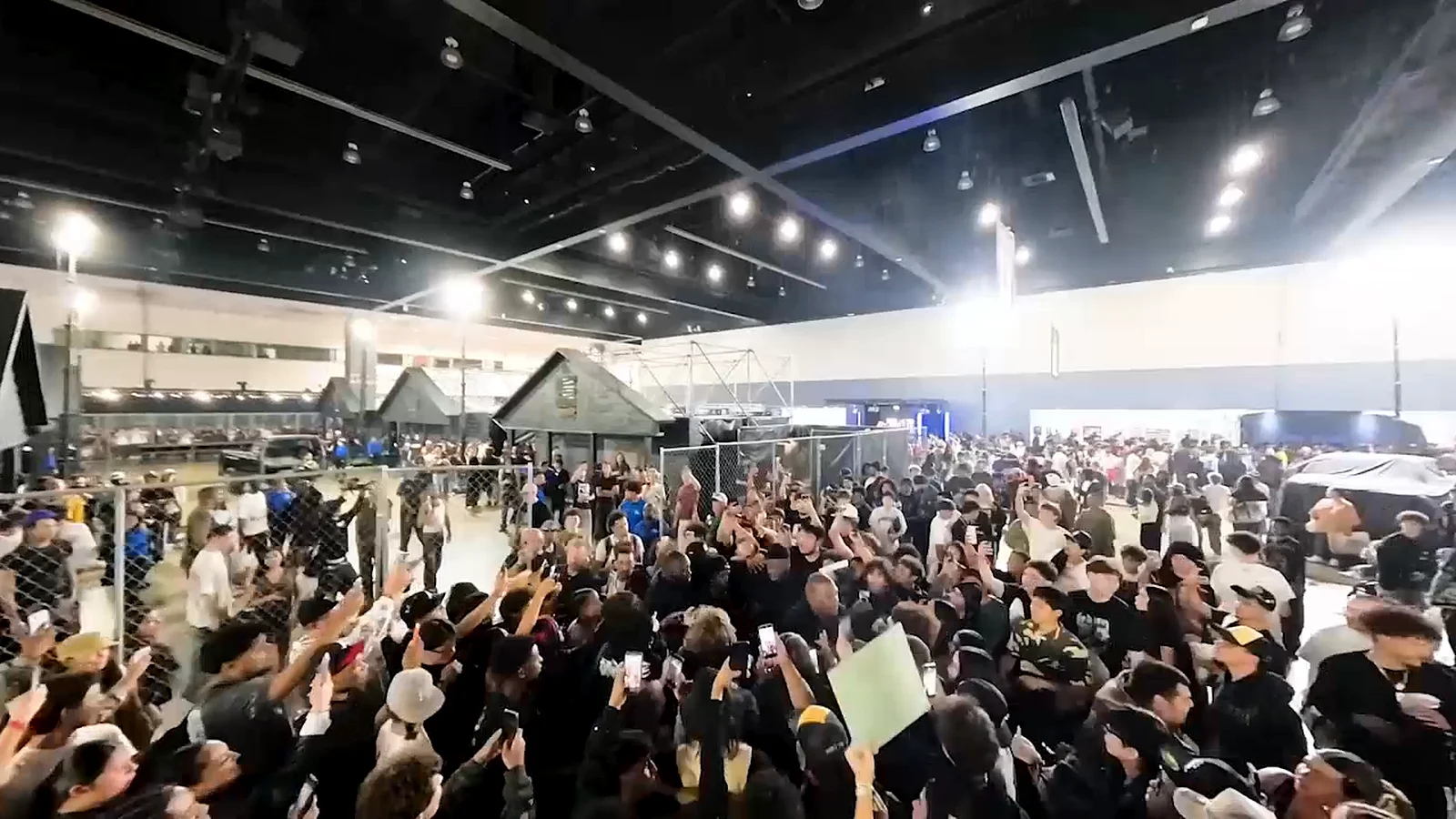 Une foule dense, téléphones levés, se presse autour d’un enclos grillagé et de petites cabanes grises dans un grand hall d’exposition éclairé par des projecteurs.