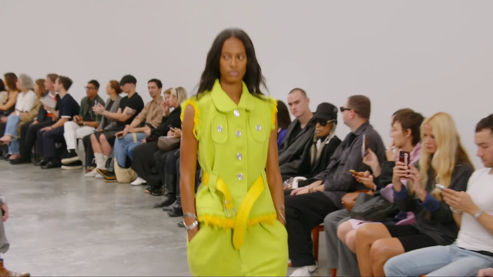 Une mannequin en combinaison sans manches vert anis, boutons métalliques et ceinture jaune, défile devant le premier rang du défilé Courrèges printemps-été 2027, dans une salle blanche au sol de béton.