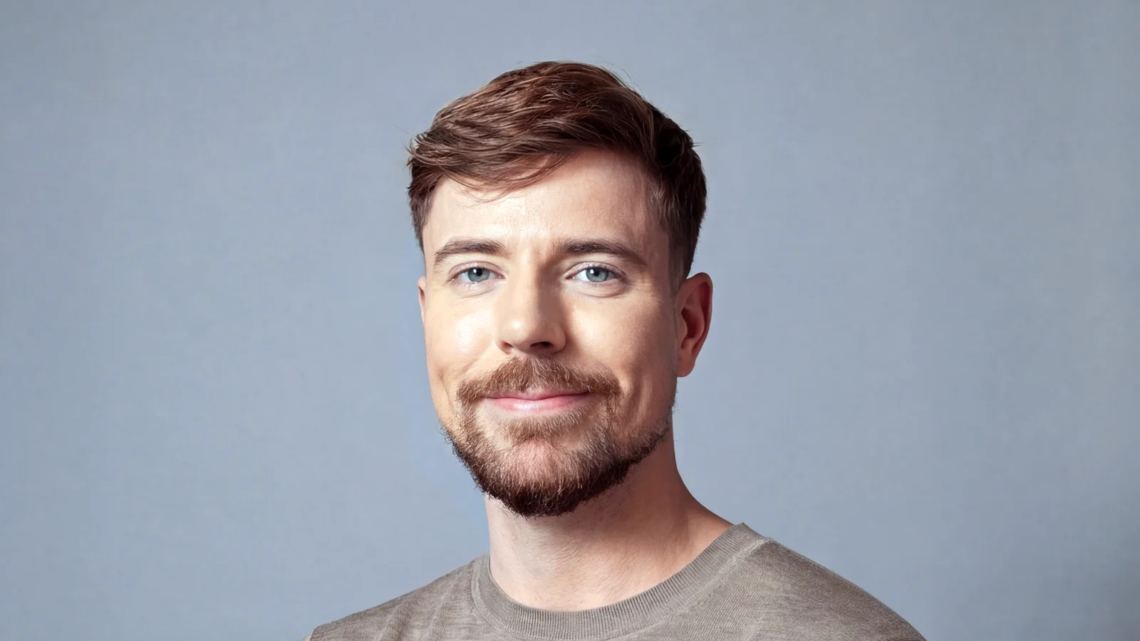 MrBeast (Jimmy Donaldson), barbe rousse et pull beige, sourit face à l’objectif devant un fond gris-bleu.