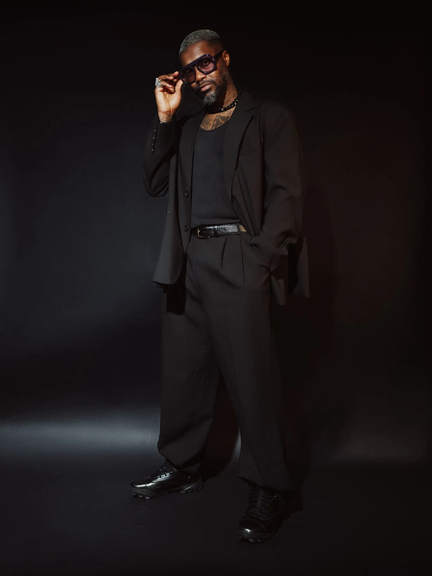 Djibril Cissé en costume noir ample et débardeur noir, debout sur fond sombre, ajuste ses lunettes violettes d’une main, l’autre dans la poche.