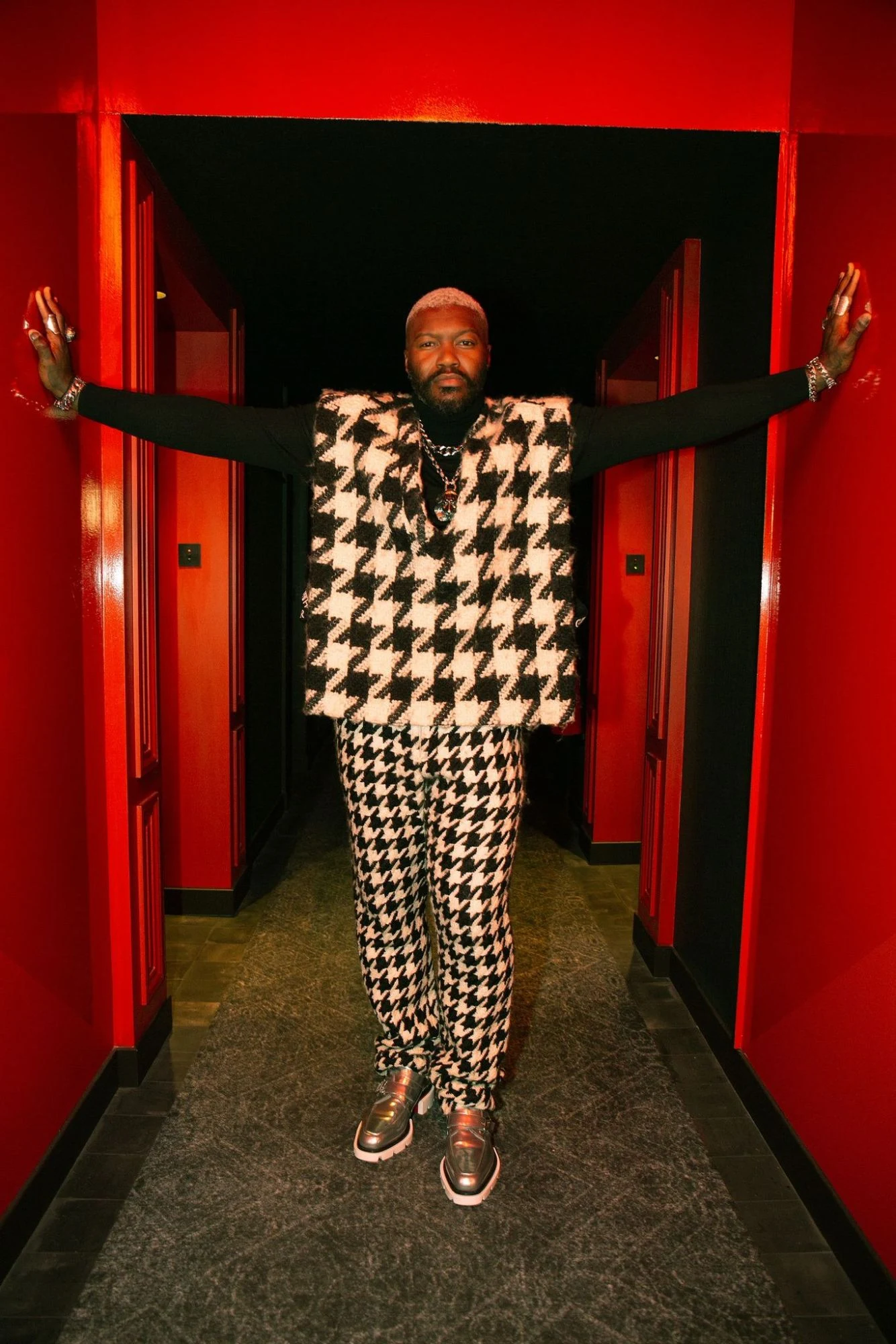 Djibril Cissé en ensemble pied-de-poule noir et blanc et mocassins argentés, bras écartés contre les murs d’un couloir rouge.