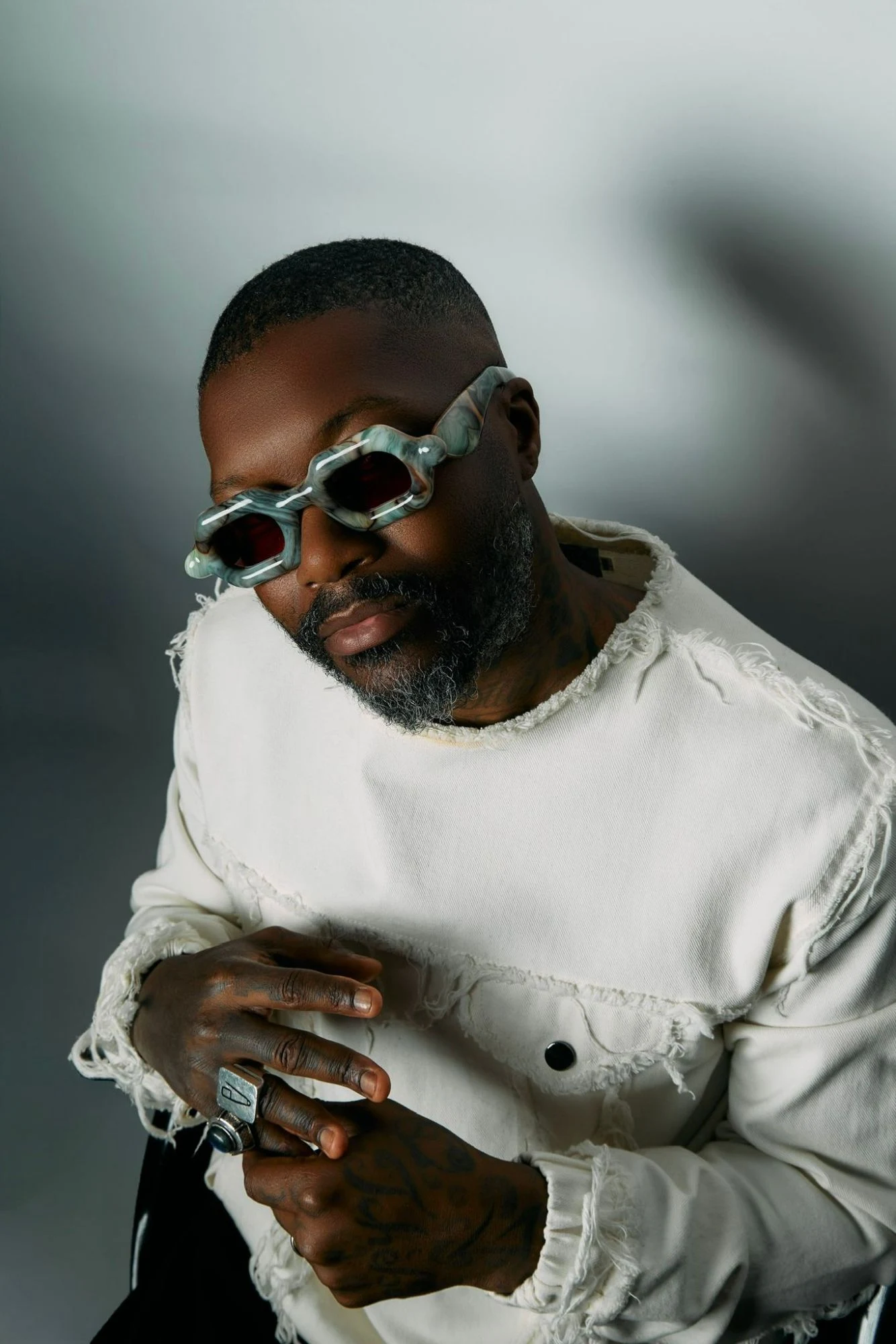 Djibril Cissé en veste blanche effilochée et lunettes à monture marbrée, les mains croisées, bagues aux doigts, sur fond gris.