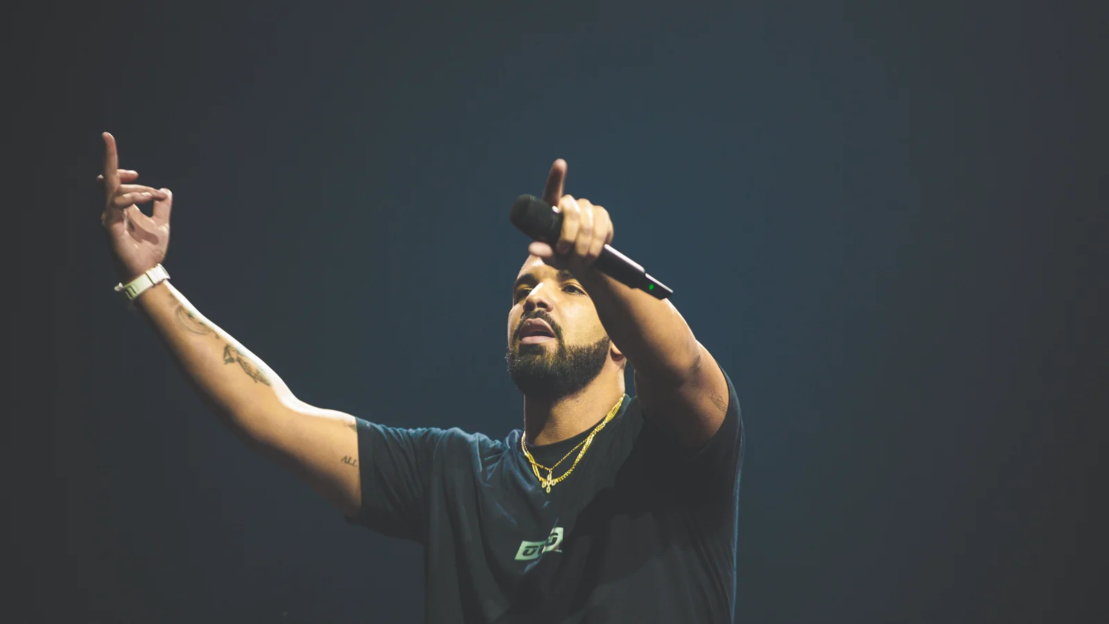 Drake sur scène, barbe et chaînes dorées, tee-shirt sombre, pointe le public de la main qui tient le micro, l’autre bras levé, sur fond bleu nuit.