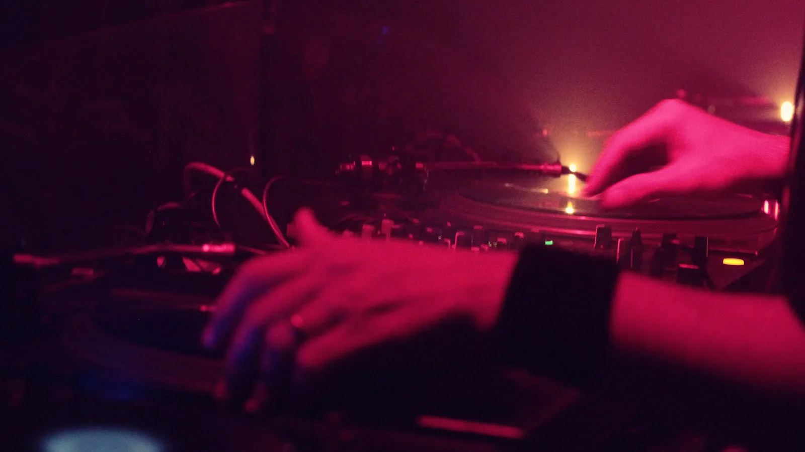 Les mains d’un DJ sur deux platines vinyles et une table de mixage, dans la lumière rouge et la fumée d’un club