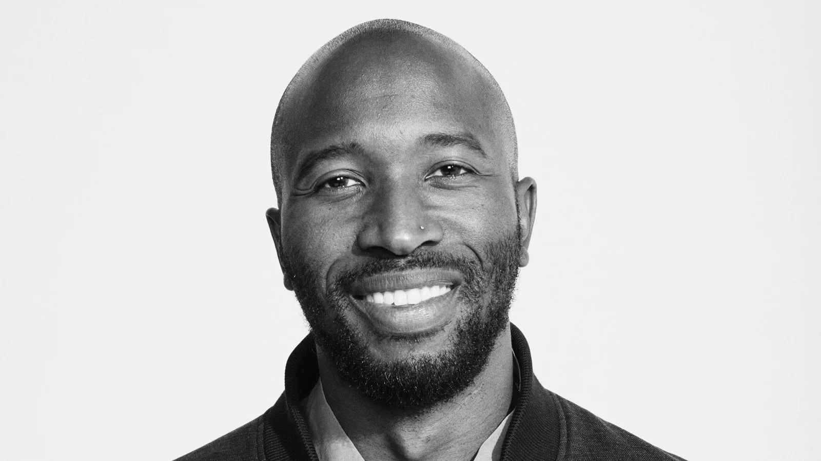 Portrait en noir et blanc d’Ezekiel Lewis, souriant, crâne rasé et barbe courte, veste sombre, sur fond blanc.