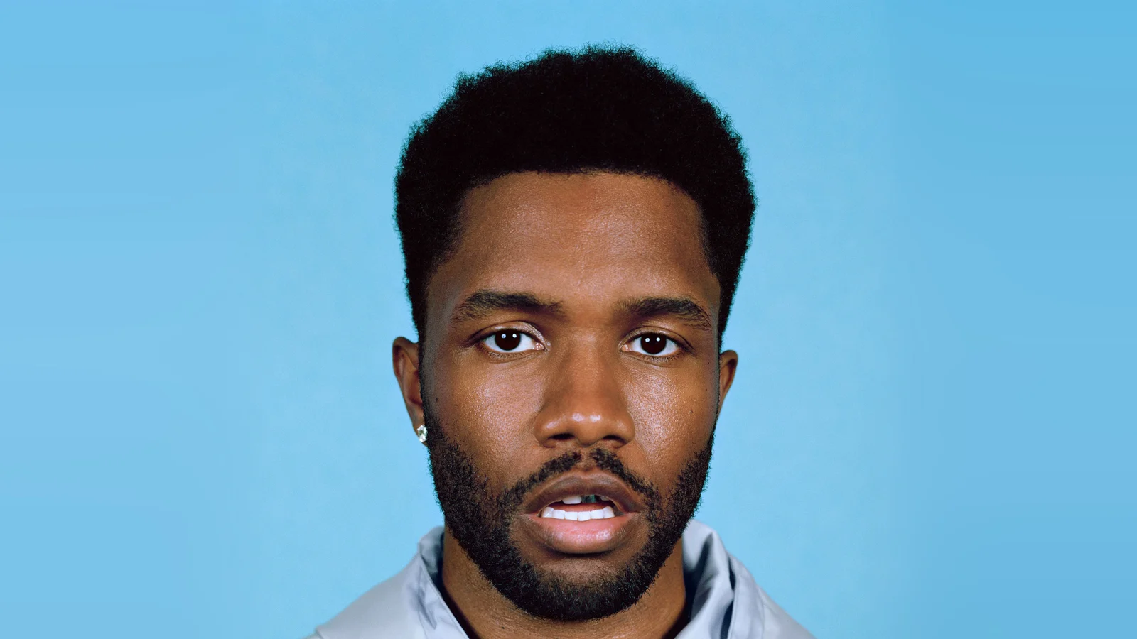 Portrait de Frank Ocean, cheveux courts et barbe, sweat à capuche gris clair, regard face à l’objectif sur un fond bleu ciel uni.