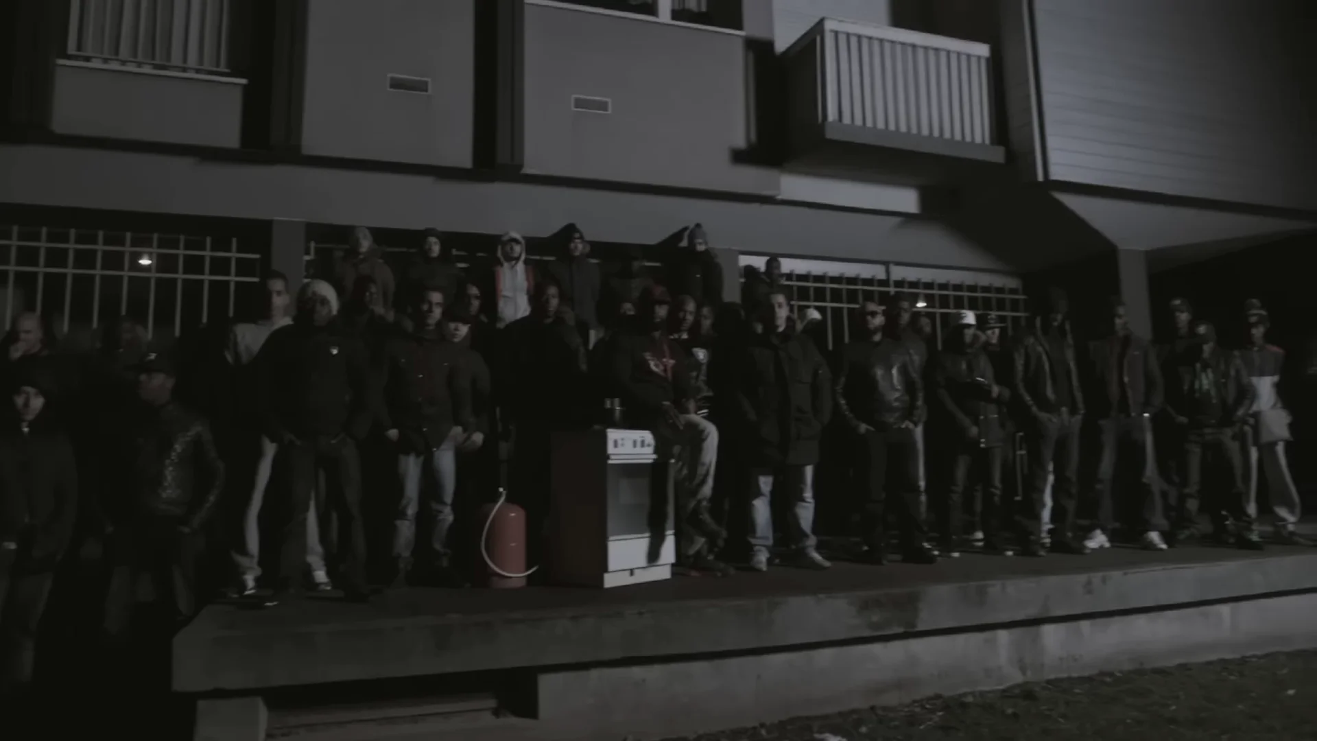 De nuit, une trentaine d’hommes alignés sur une dalle au pied d’un immeuble, sous une lumière blafarde.