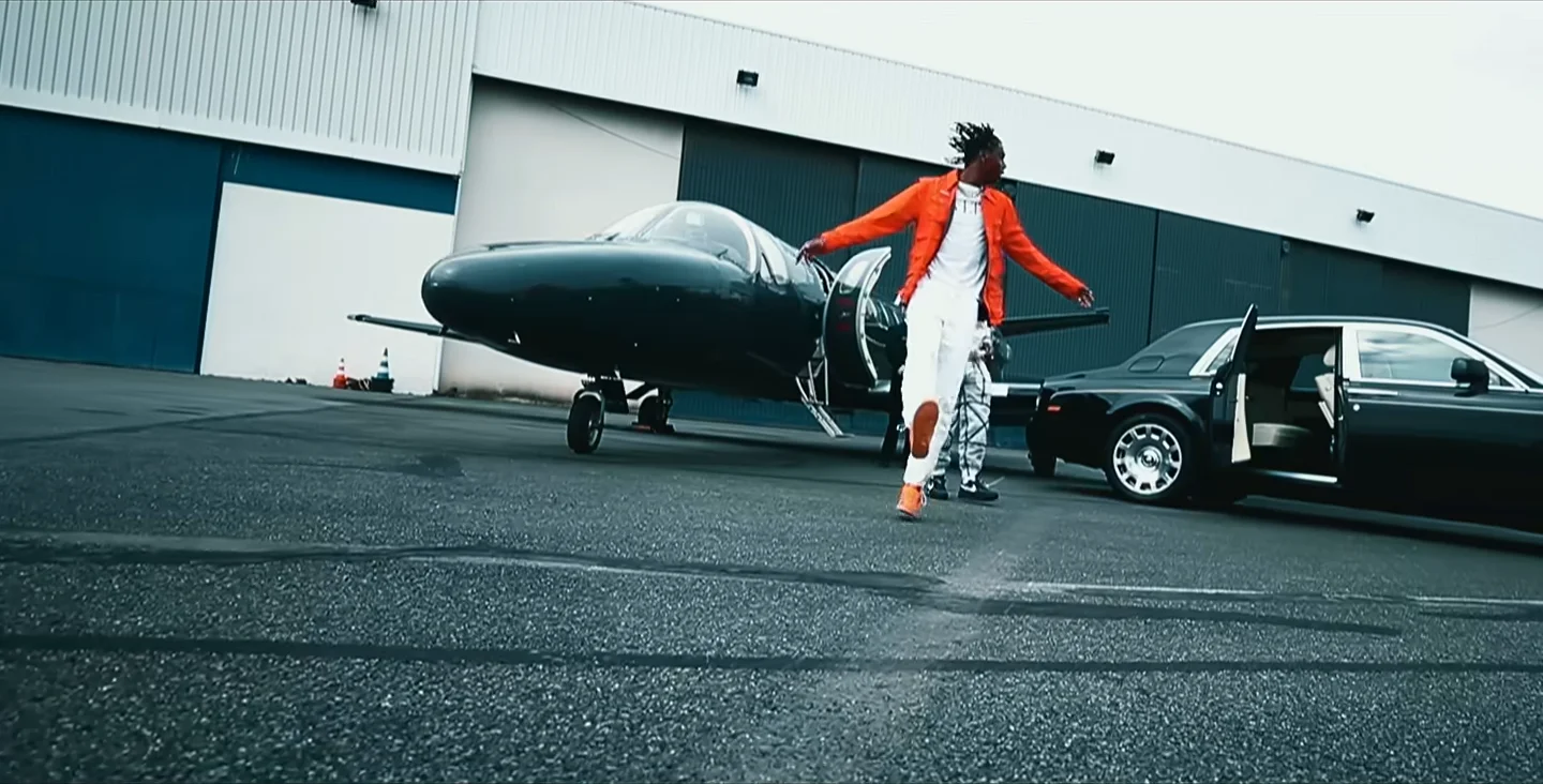 Un rappeur en veste orange et pantalon blanc danse sur le tarmac, entre un jet privé noir et une berline noire portière ouverte, devant un hangar.