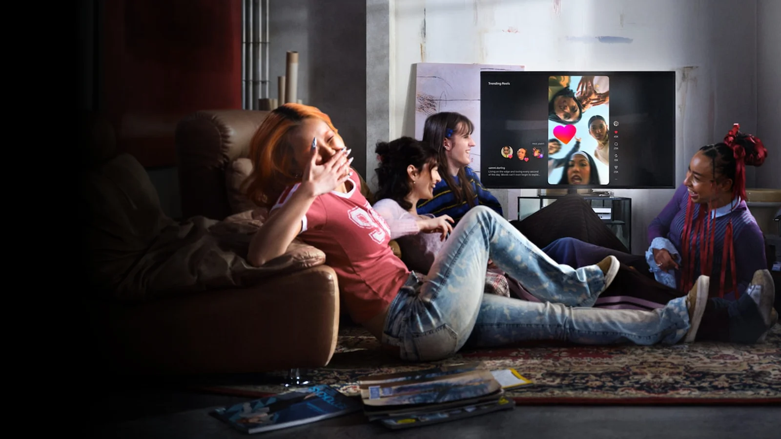 Quatre jeunes femmes rient dans un salon, assises sur un canapé et sur un tapis, devant un téléviseur qui affiche des reels Instagram.