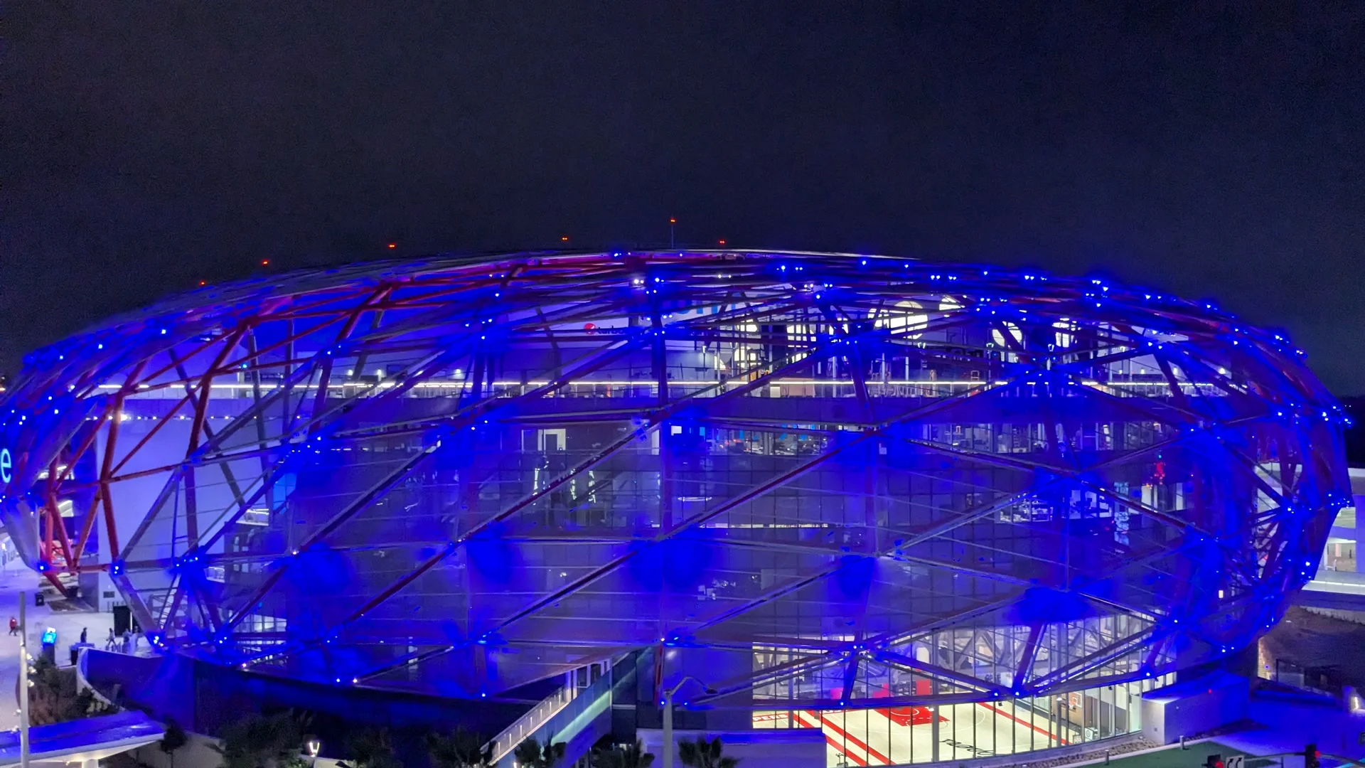 L’Intuit Dome, la salle des Los Angeles Clippers à Inglewood, de nuit : la structure en treillis de la façade, éclairée en bleu et rouge, enveloppe les étages vitrés.