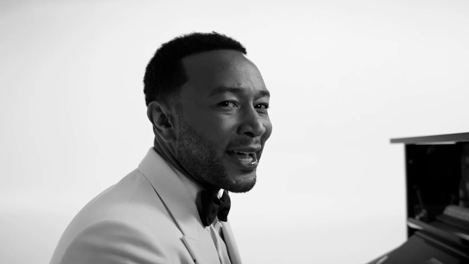 John Legend, en smoking blanc et nœud papillon noir, chante en souriant devant un piano droit, sur fond blanc, en noir et blanc.