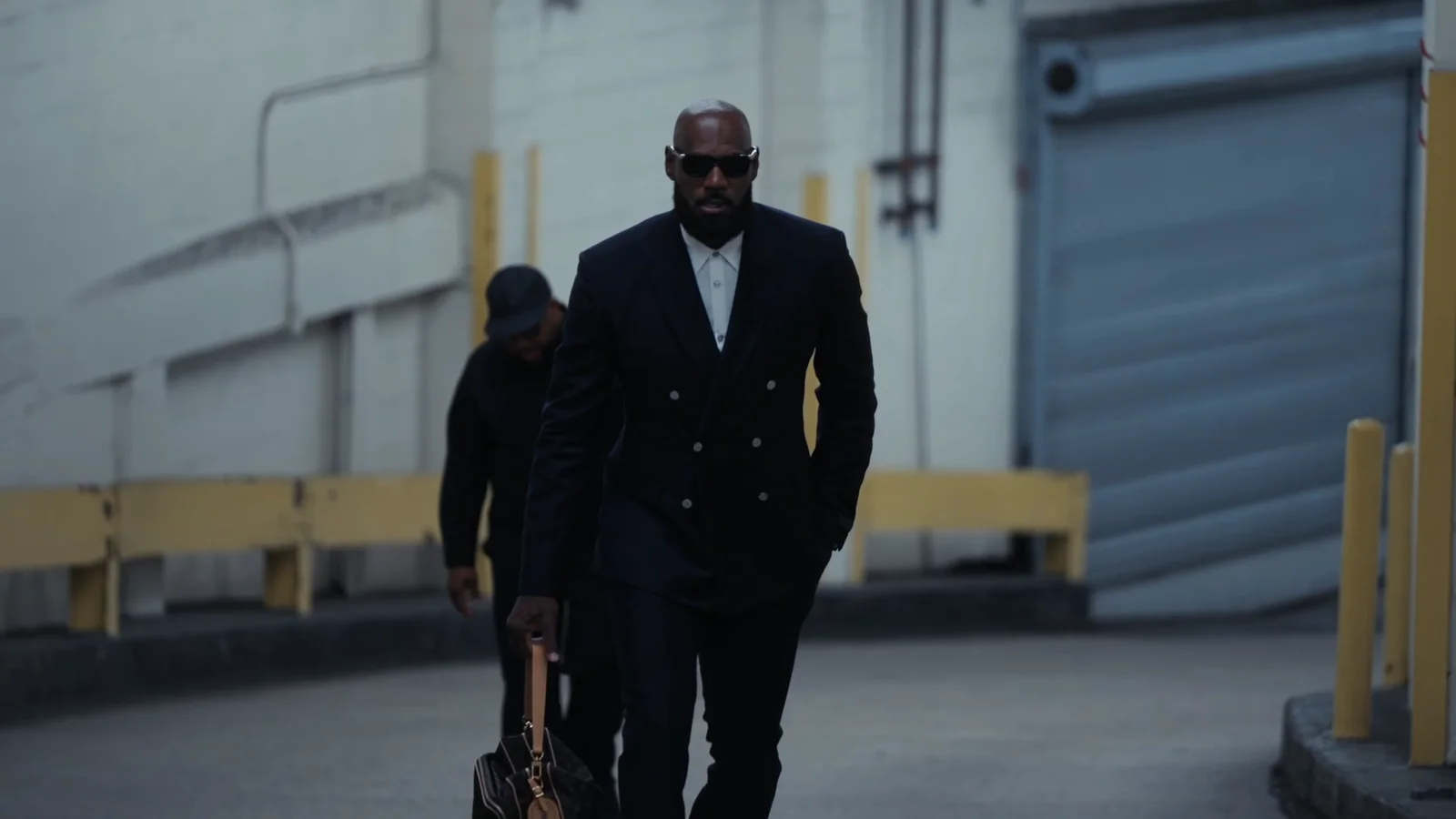 LeBron James, en costume croisé noir et lunettes de soleil, marche dans un couloir de service aux murs blancs, un sac à la main.