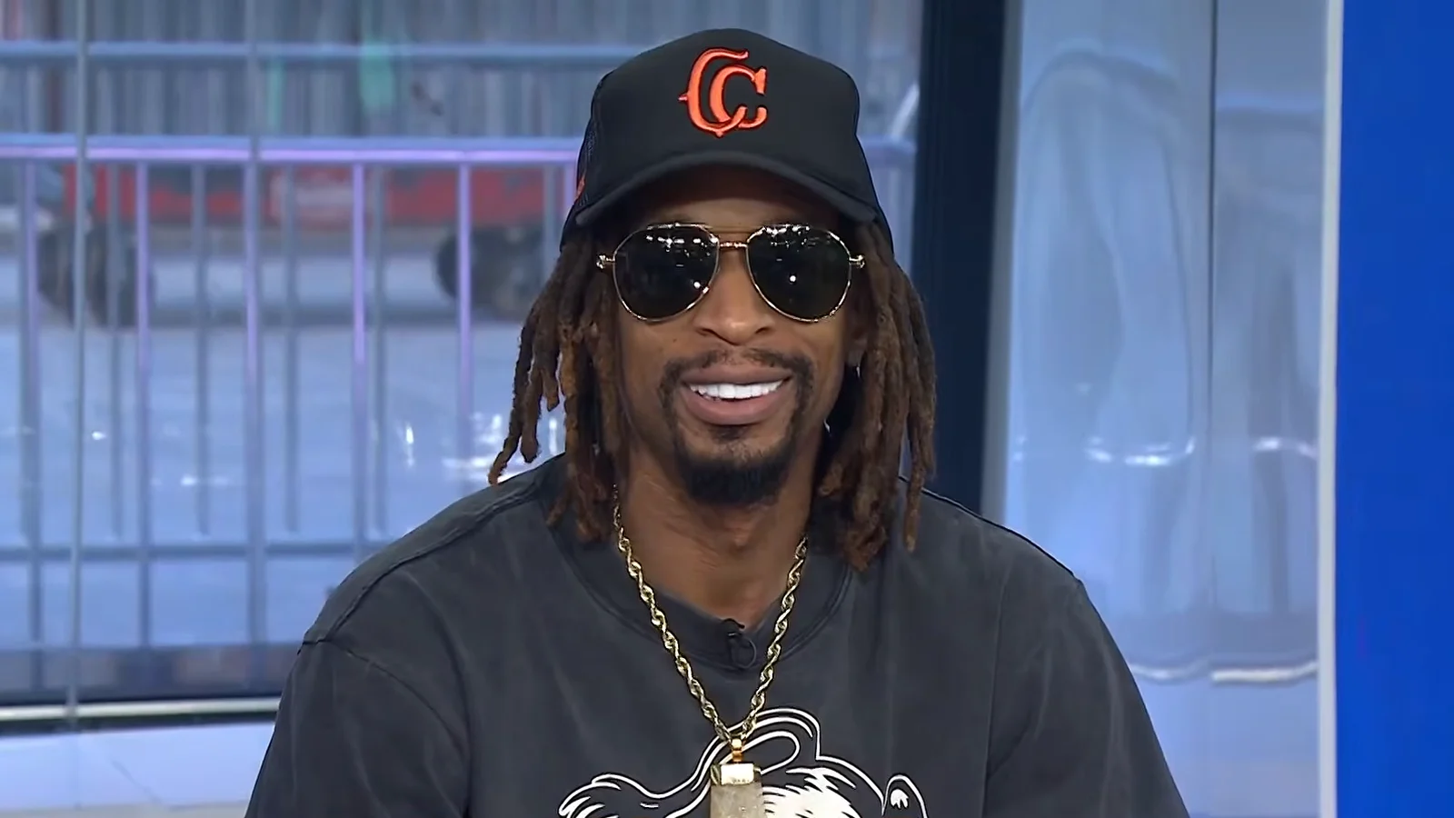 Lil Jon, casquette noire brodée, lunettes de soleil aviateur et dreadlocks, sourit en tee-shirt gris délavé et chaîne dorée sur un plateau de télévision.