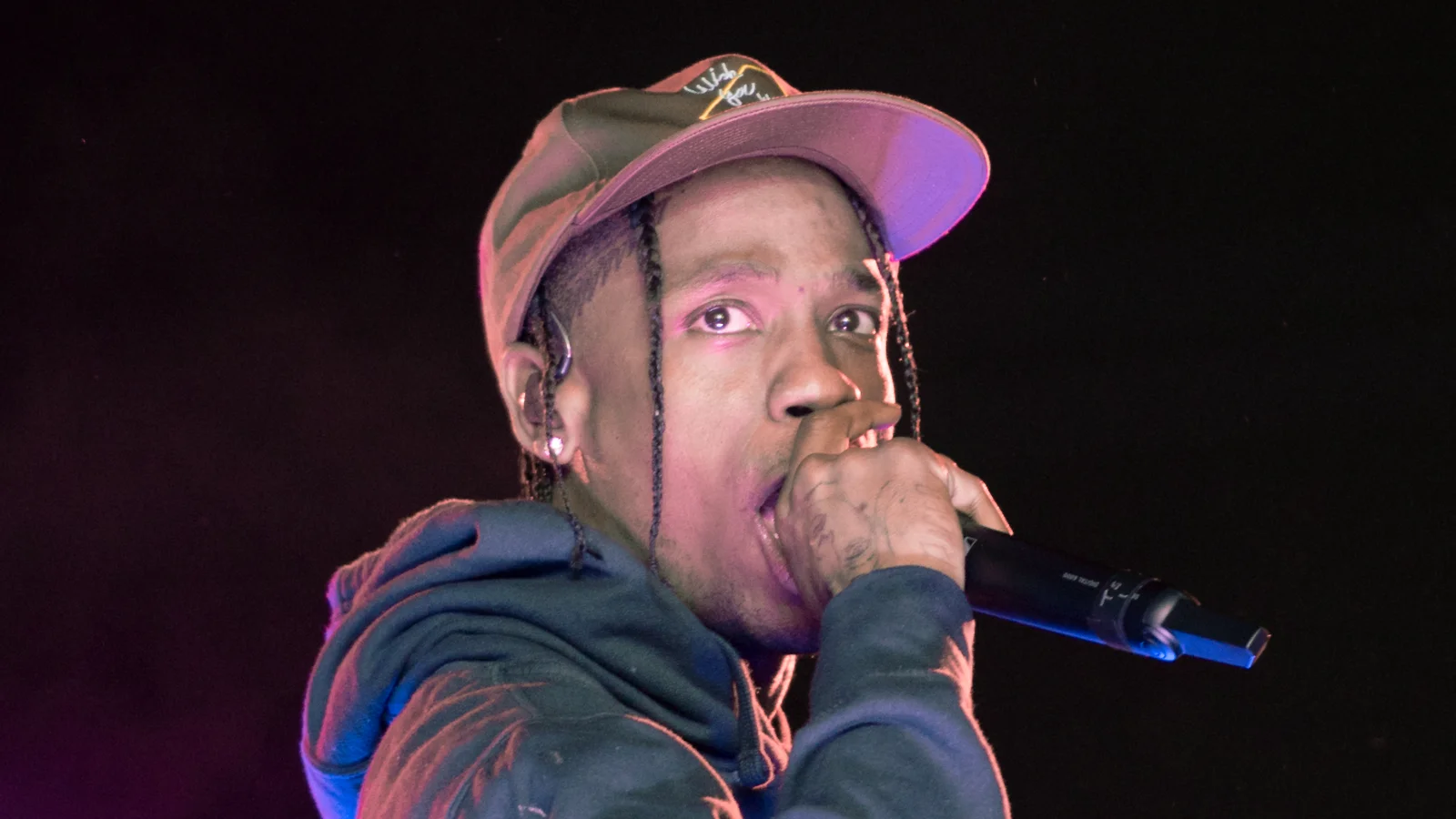 Travis Scott sur scène, casquette et sweat à capuche sombre, chante en tenant le micro contre sa bouche, sur fond noir.