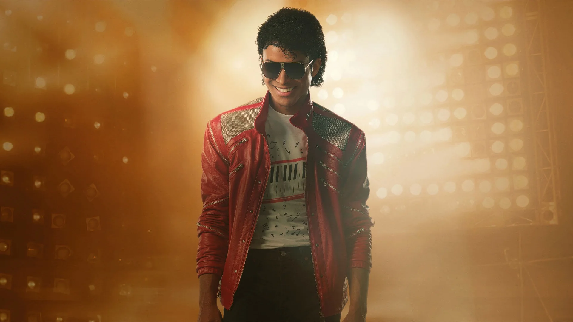 Jaafar Jackson, en veste rouge et lunettes de soleil, sourit devant un mur de projecteurs dans le film « Michael ».