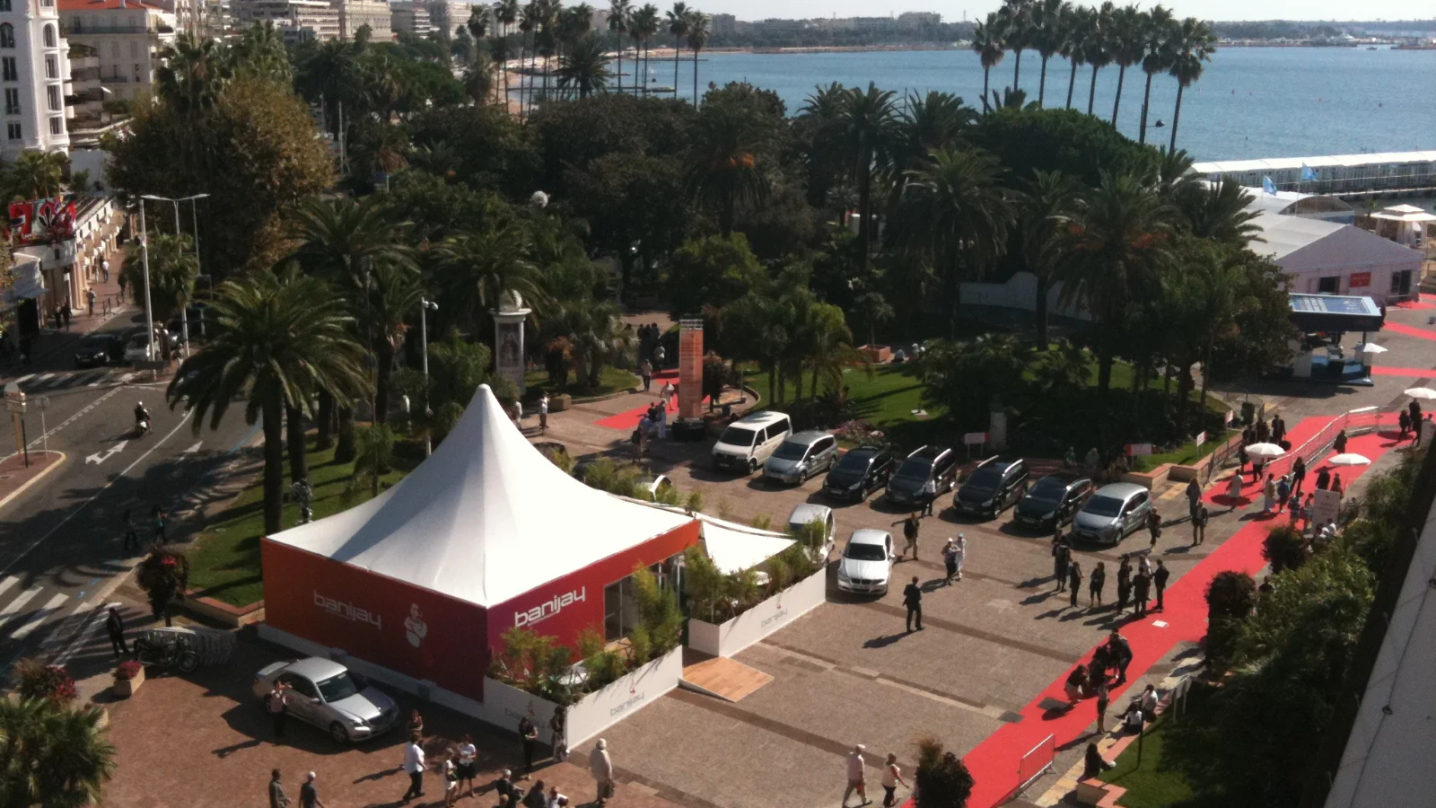 Vue plongeante sur les abords du Palais des Festivals pendant le MIPCOM 2010 : une tente blanche et rouge d'exposant, des voitures garées, des palmiers et la baie de Cannes en arrière-plan