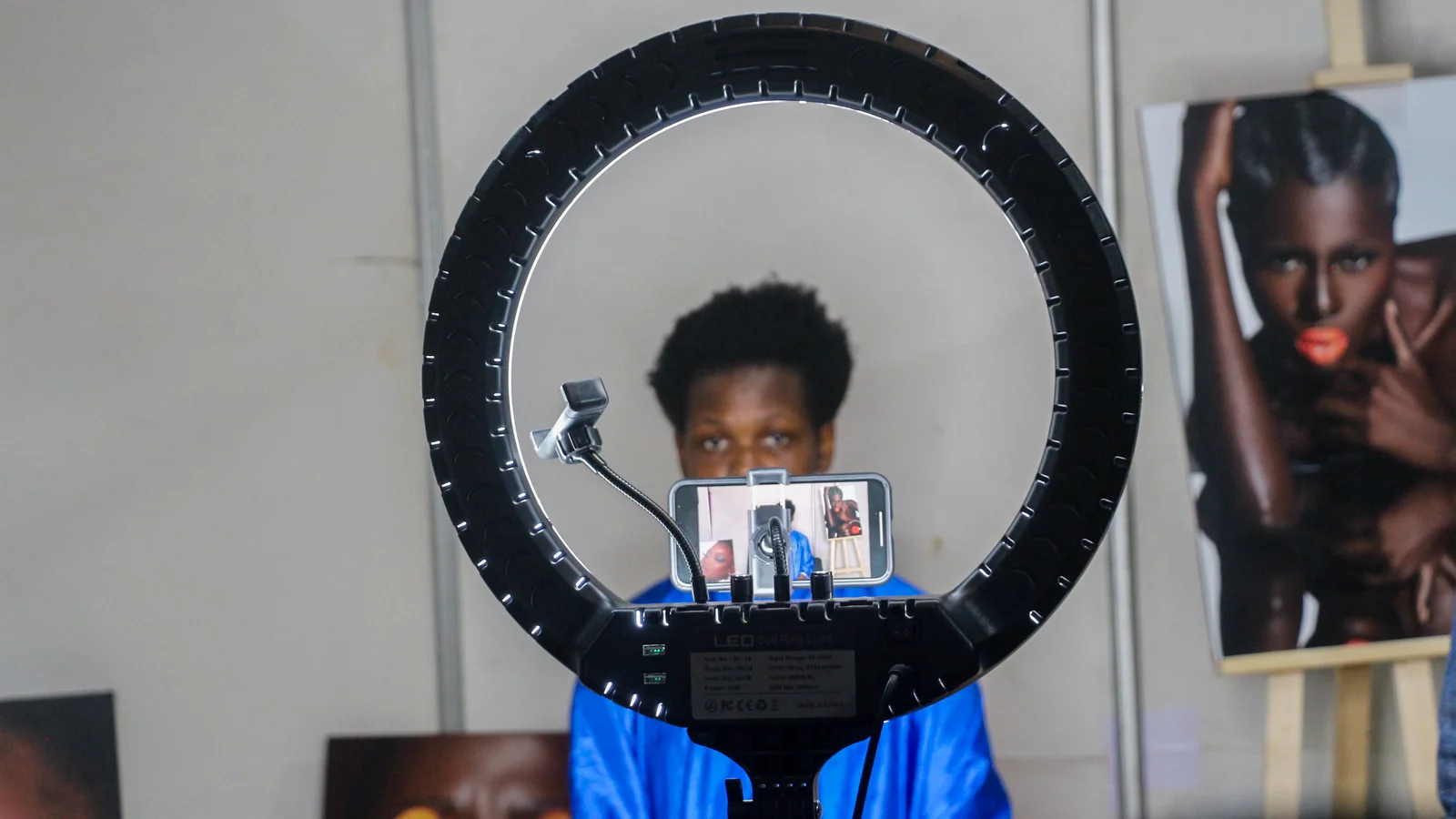 Un jeune créateur filme une vidéo face à son smartphone, fixé au centre d’un grand anneau lumineux noir, devant des tirages photo posés sur des chevalets.