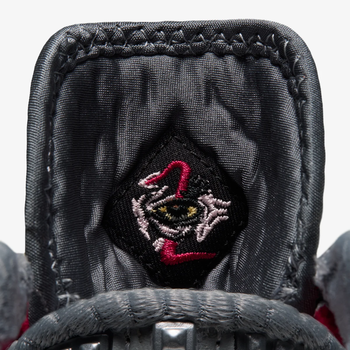 Gros plan sur la languette grise de la LeBron XXIV « Storm Drain » : un écusson noir brodé d’un œil jaune dans un tourbillon blanc et rouge.