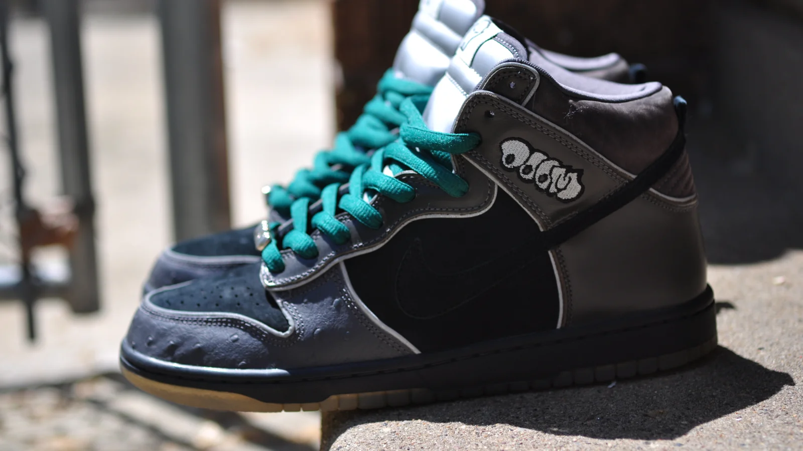 Paire de Nike SB Dunk High grises et noires à lacets verts, posée sur un muret en plein soleil.