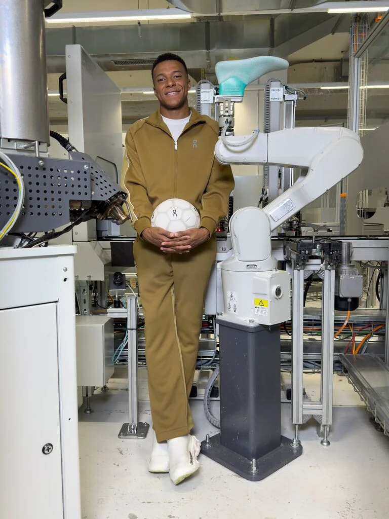 Kylian Mbappé debout avec un ballon On dans le laboratoire de la marque.