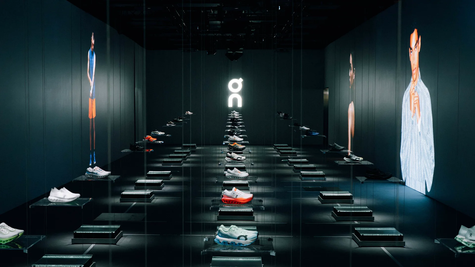 Salle d’exposition plongée dans la pénombre lors de l’Investor Day d’On : des dizaines de chaussures On alignées sur des socles éclairés, le logo On lumineux au fond et, sur les murs, des écrans géants montrant des athlètes et des mannequins.