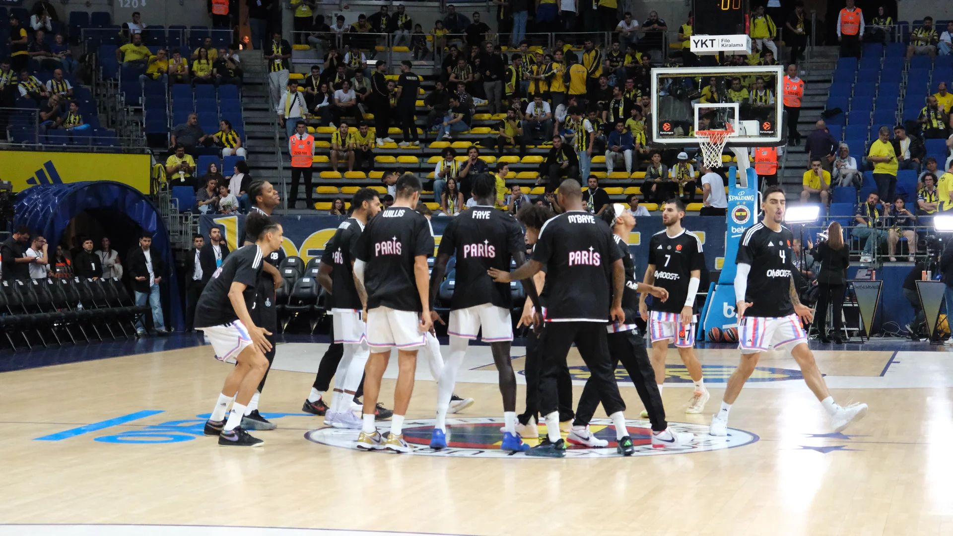 Les joueurs de Paris Basketball, en tee-shirts noirs floqués « Paris », réunis au centre du parquet avant un match d’EuroLeague à Istanbul, devant des tribunes de supporters du Fenerbahçe en jaune.