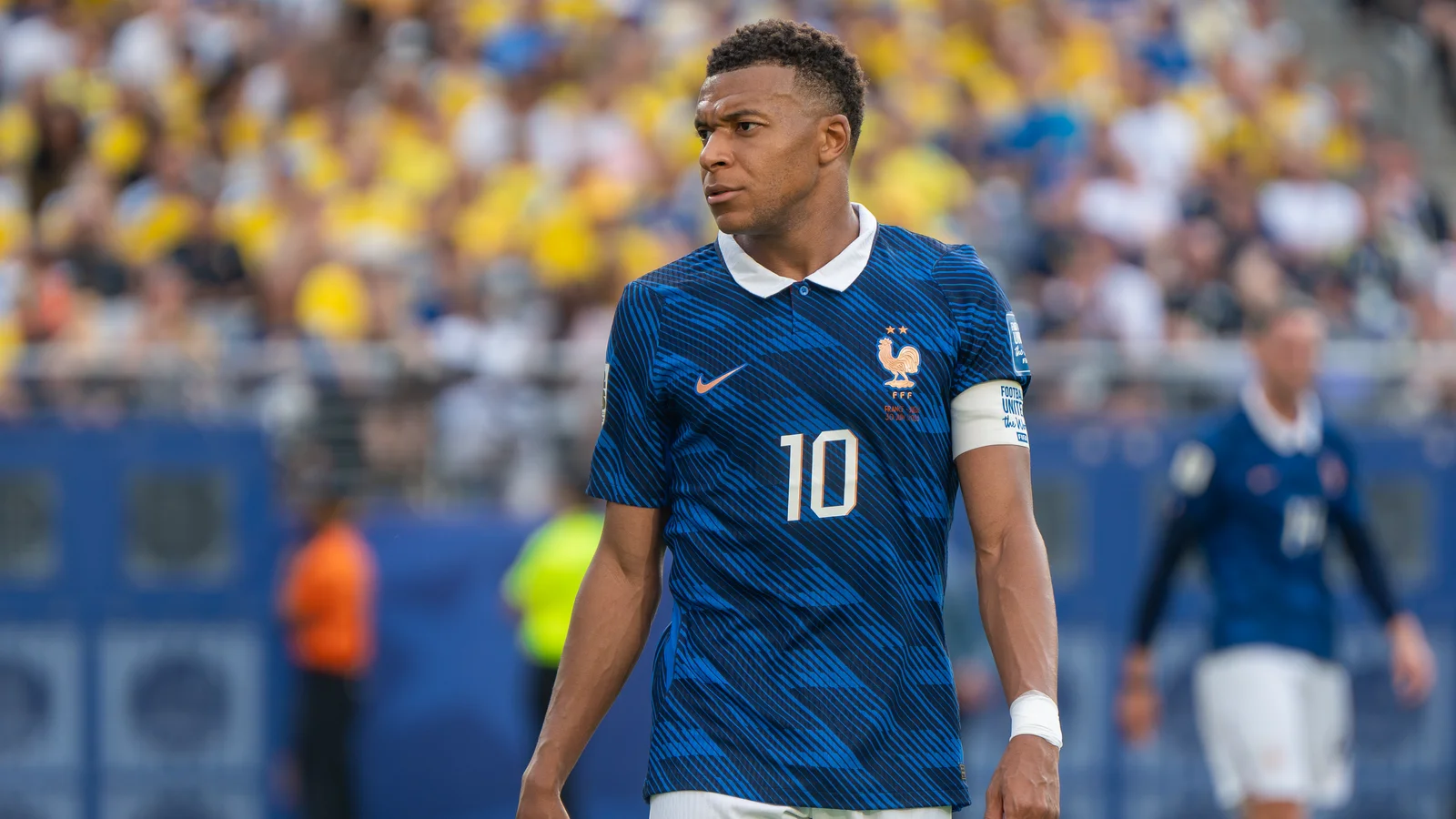 Kylian Mbappé, maillot bleu numéro 10 et brassard de capitaine de l’équipe de France, regarde sur le côté pendant le match France-Suède de la Coupe du monde 2026, devant des tribunes jaunes.