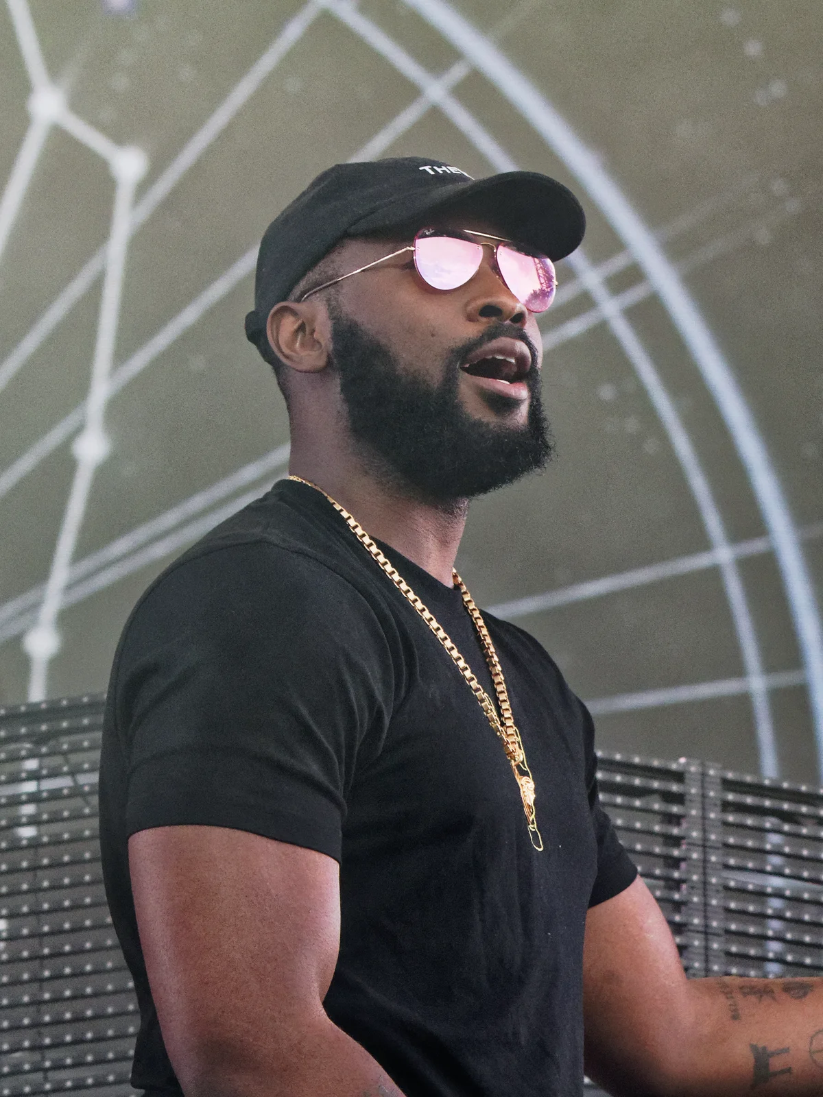 Damso en tee-shirt noir, casquette noire et lunettes rosées, une chaîne dorée autour du cou.