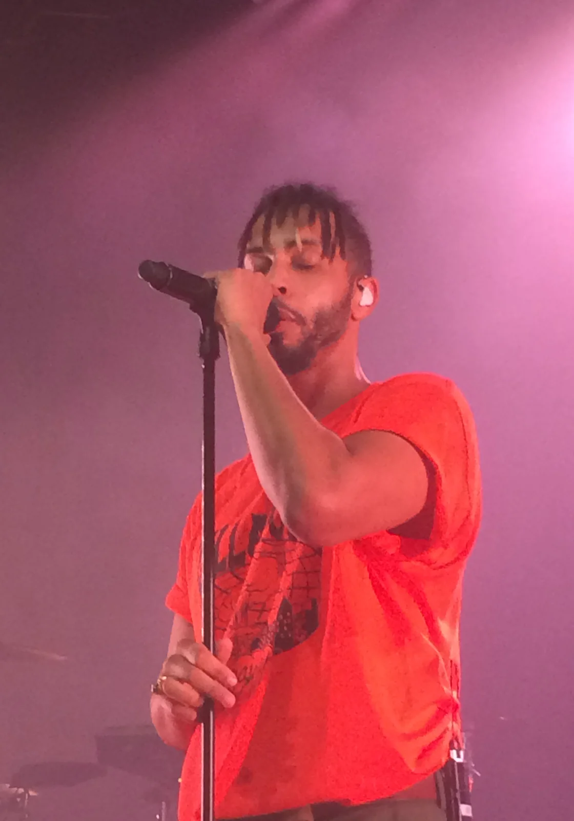 Disiz en tee-shirt rouge, la main sur le pied de micro, sous une lumière rose.