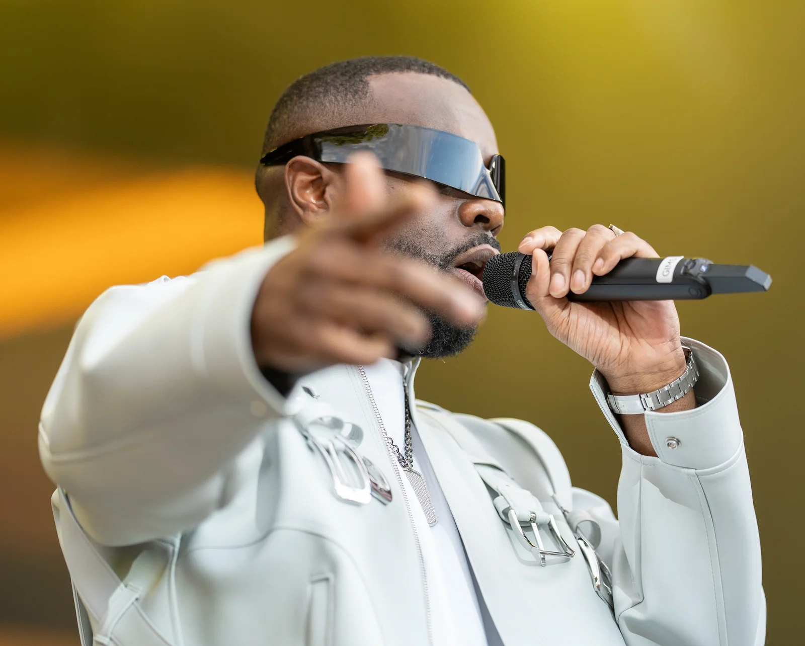Gims en veste blanche et lunettes de soleil, le bras tendu vers le public, micro à la main.