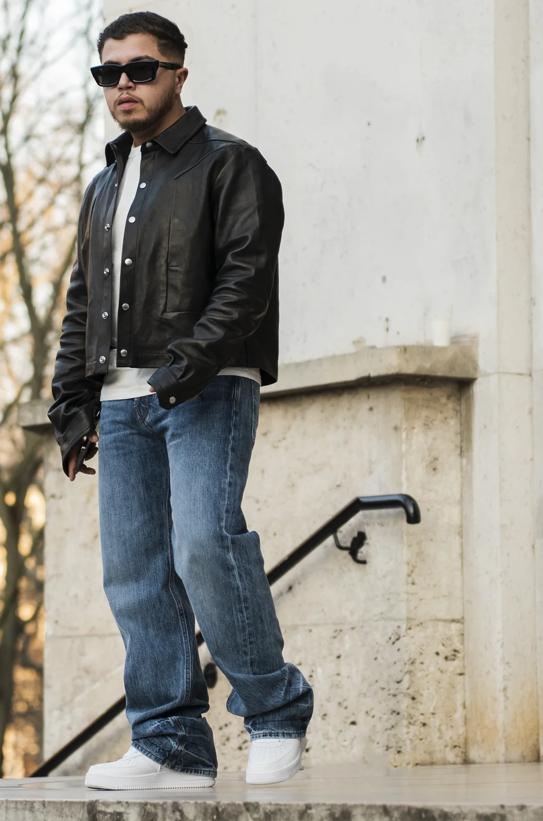 Hamza en blouson de cuir noir, jean large et lunettes de soleil, debout sur des marches de pierre.