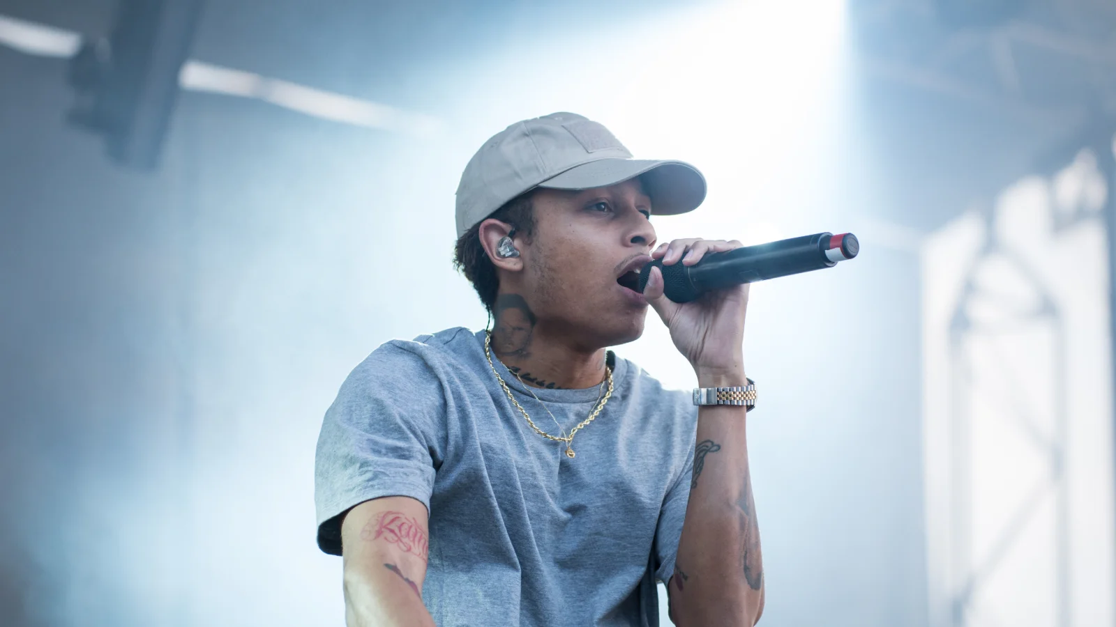 Le rappeur Joke, casquette beige et t-shirt gris, chante au micro sur une scène de festival baignée d'une lumière blanche bleutée